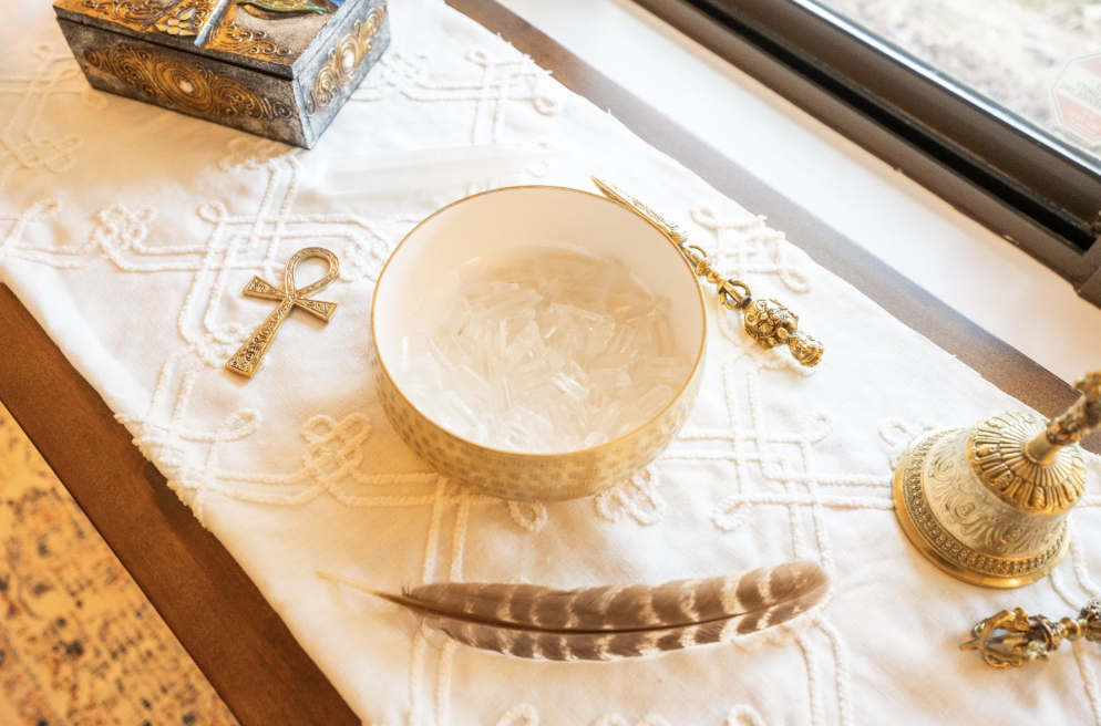 Decorative altar table with gold and white items, including an ankh pendant, a feather, a bowl with clear crystals, a candlestick, and a box, near a window.