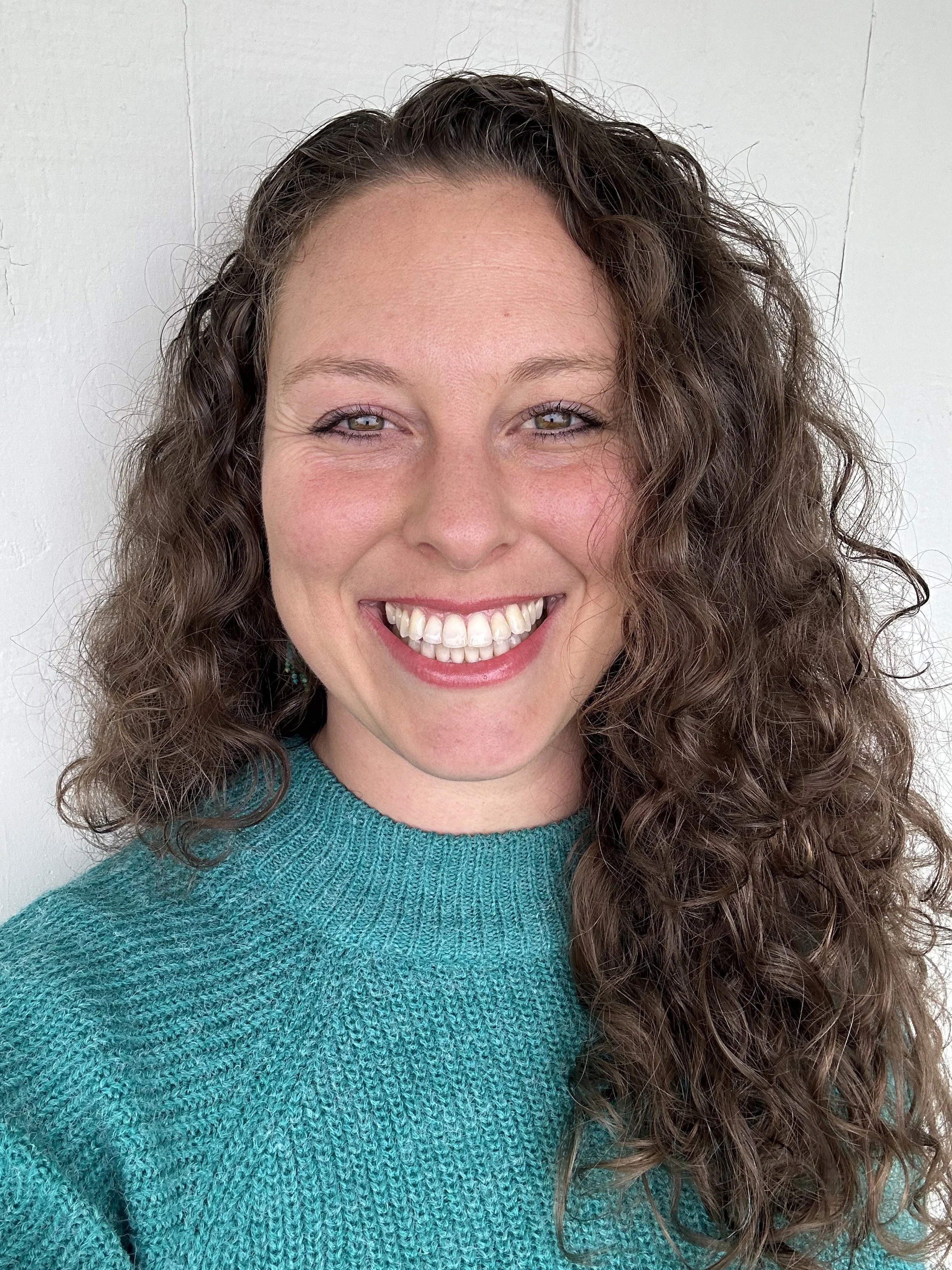 A woman with curly brown hair, light eyes, and a wide smile, wearing a teal knit sweater, standing against a plain white background.