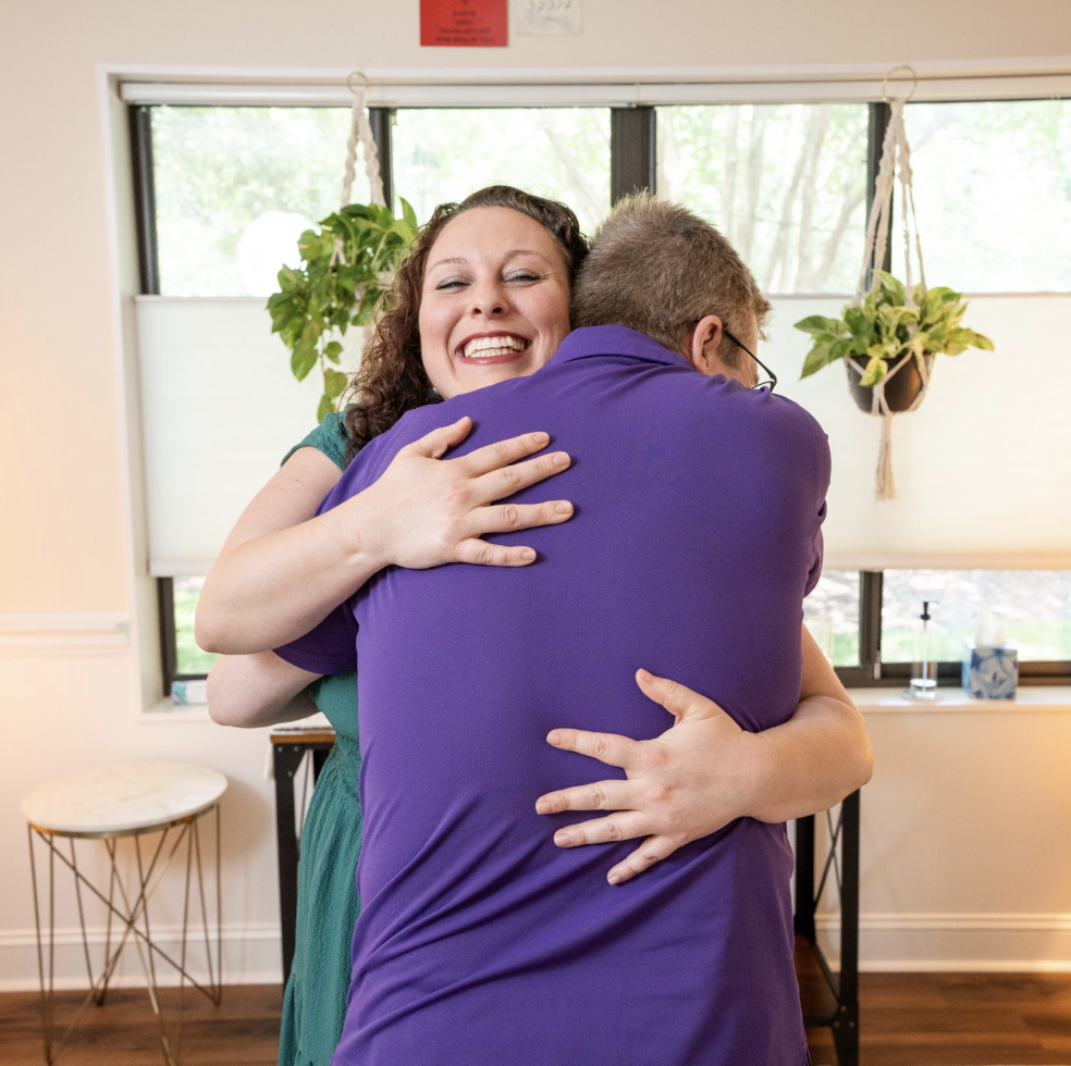 A woman with curly hair smiling and hugging a man with glasses from behind in a room with blinds, hanging plants, and a small table.