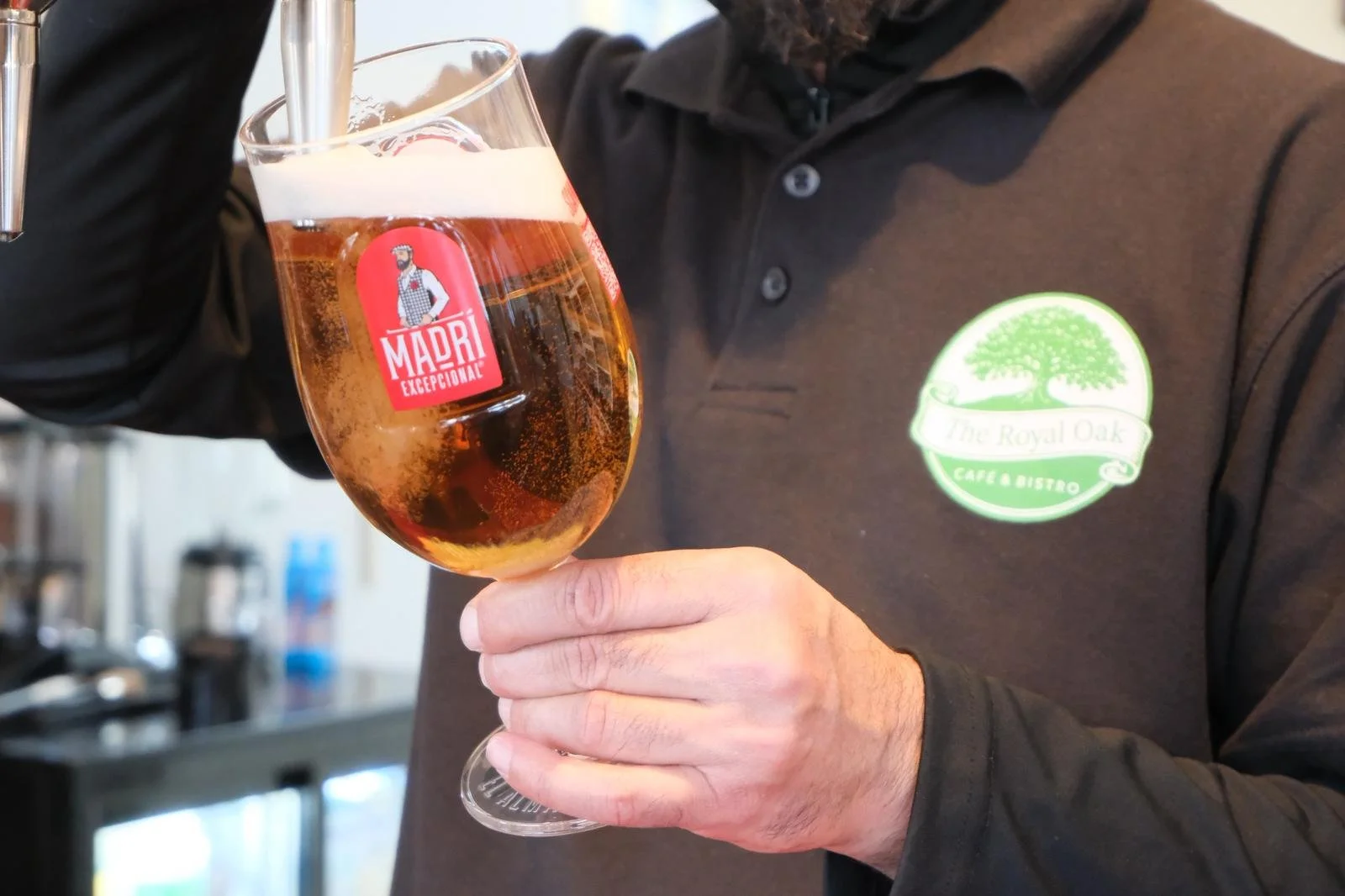 A person wearing a black shirt with a green and white logo holds a glass of beer with a stem, with foam on top. The beer glass has a red sticker with a man in a chef's hat and apron and the text 'MADRI EXCEPCIONAL'. The background is blurred.