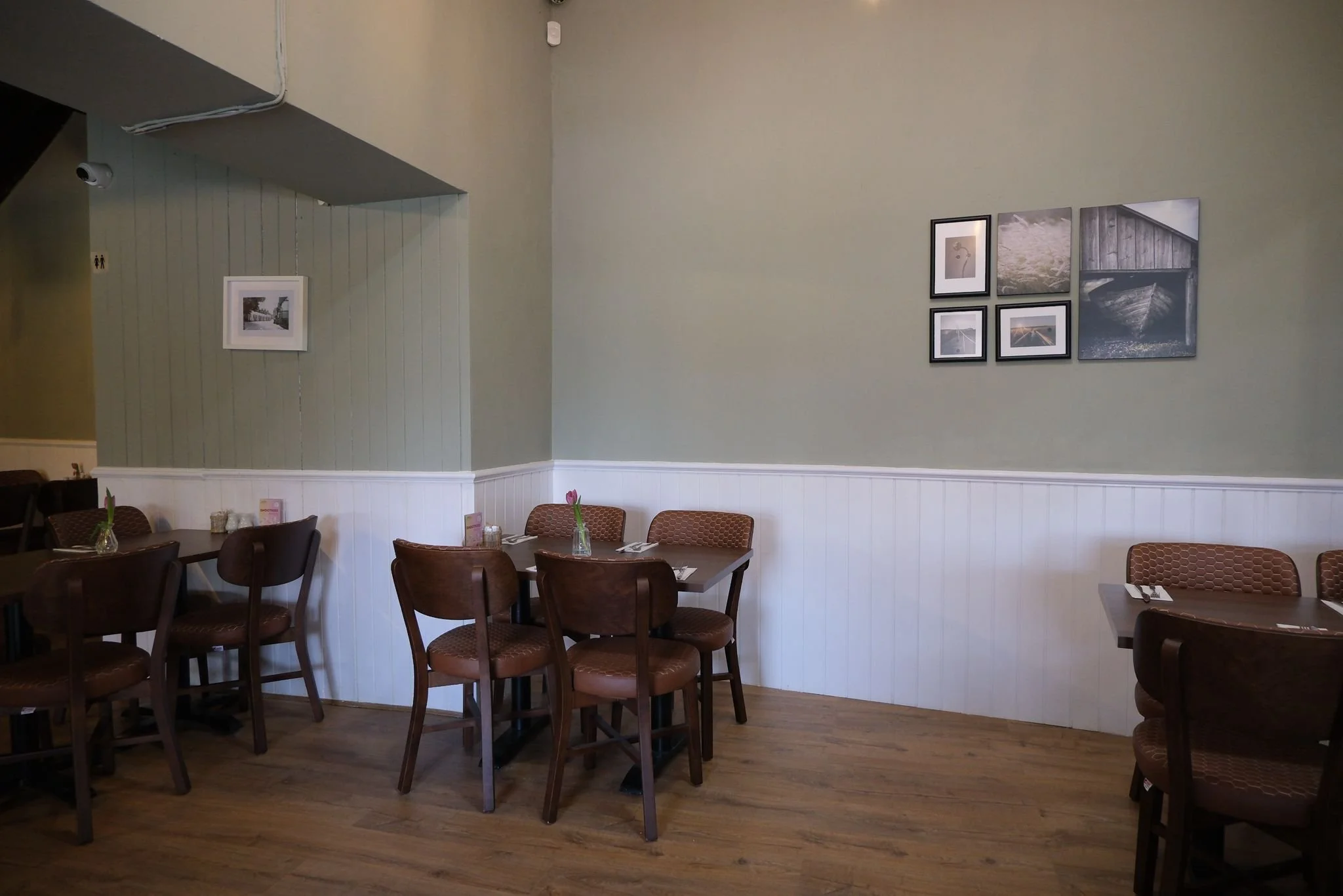 Empty restaurant with wooden chairs and tables, small vases with pink tulips, and framed photographs on a light green wall with white beadboard paneling.