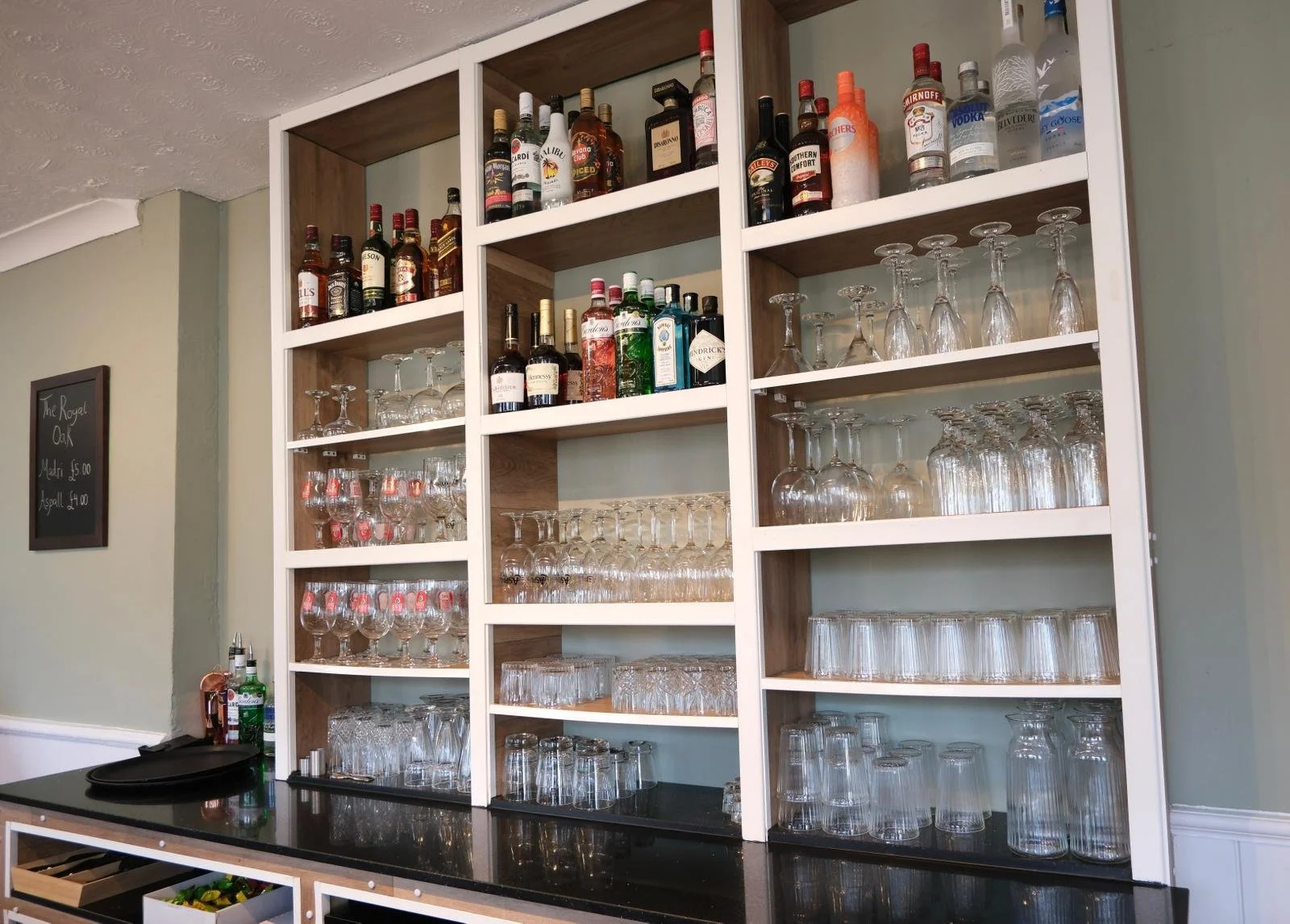 Open wooden shelving unit filled with various bottles of liquor, wine glasses, and upside-down glassware in a bar setting.