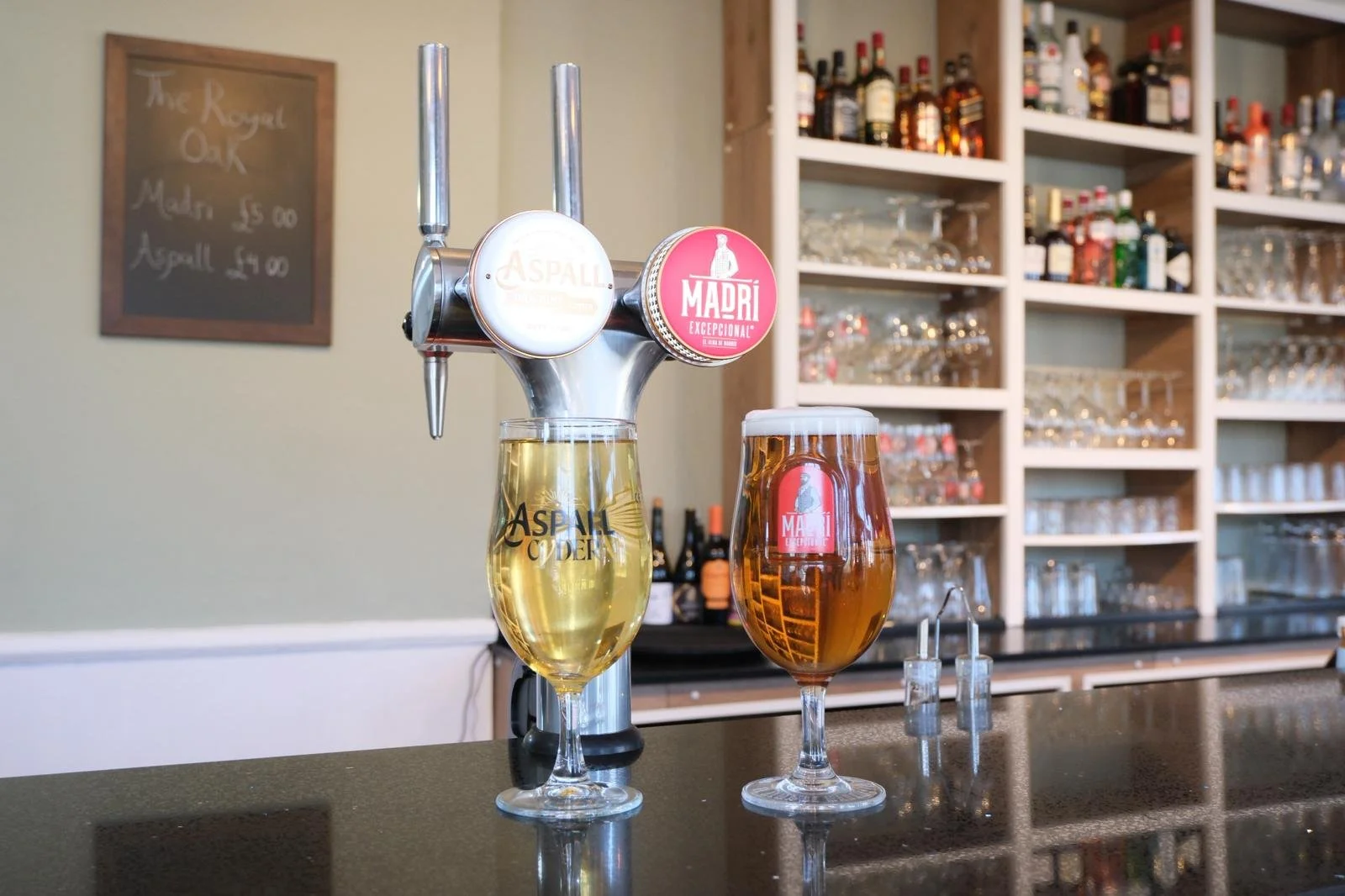 Two glasses of beer, one pale and one darker, on a bar counter with beer taps in the background.