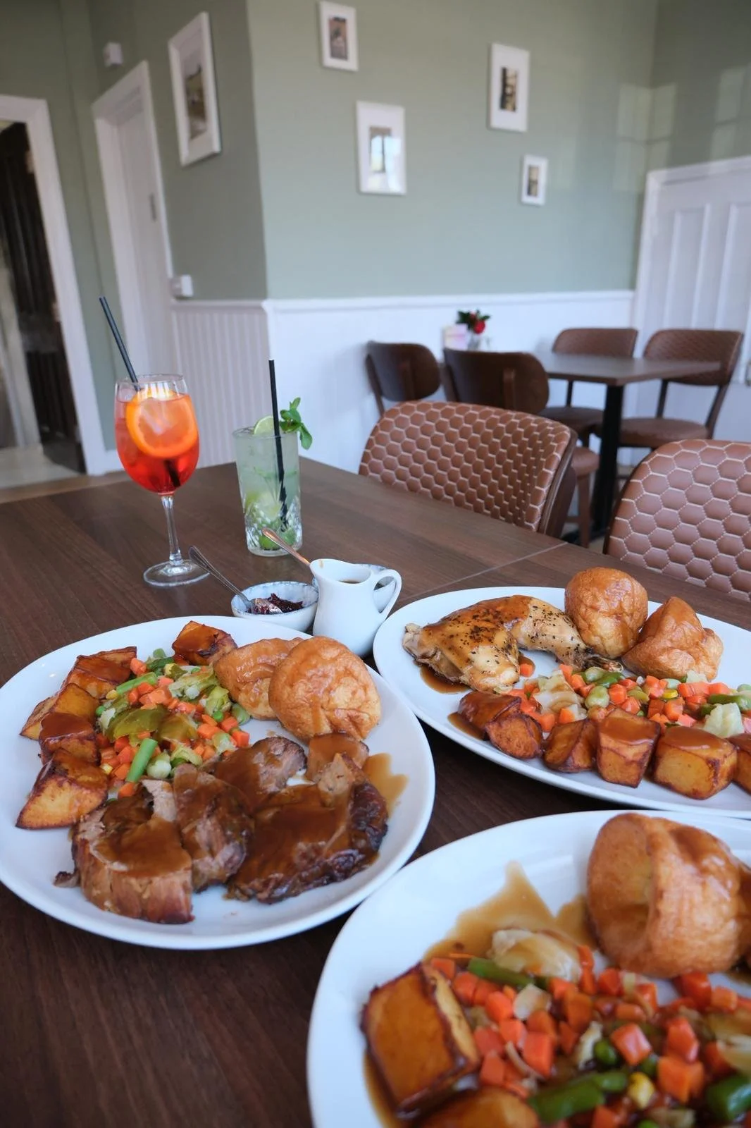 Three plates of roast dinner with vegetables, potatoes, and gravy on a wooden table in a restaurant or cafe setting, with drinks in the background.