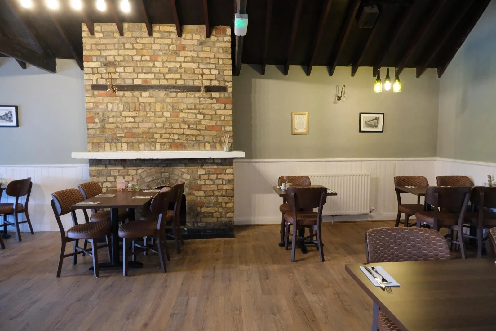Interior of a cozy restaurant with wooden floors, a brick fireplace, and tables with brown chairs. Decorative framed pictures hang on the walls, and the lighting is warm, with brass and green hanging light fixtures.