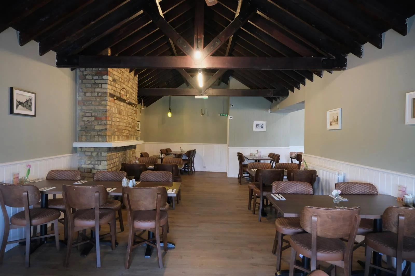 Interior of a restaurant with wooden ceiling beams, brick chimney, and framed pictures on light-colored walls. Several tables are set with placemats, utensils, and small flower vases.