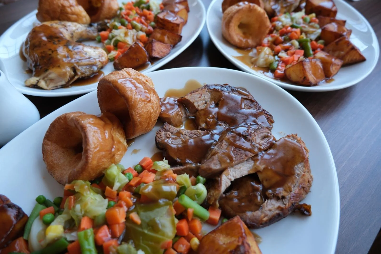 Three plates of food featuring roasted meat with gravy, fried potatoes, vegetable medley, and Yorkshire pudding.