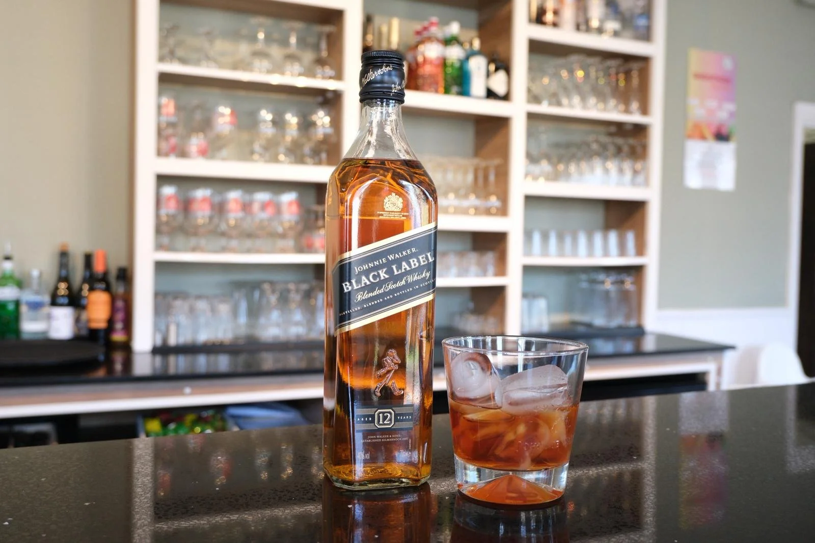 A bottle of Johnnie Walker Black Label single malt scotch whiskey with a glass of whiskey and ice on a bar counter, with glass shelves of various liquor bottles and glassware in the background.