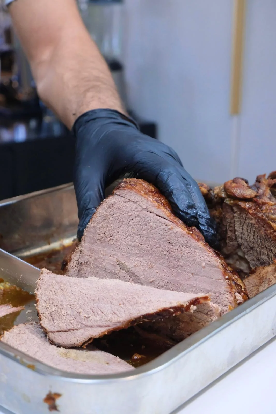 Person wearing a black glove slicing a large piece of cooked beef brisket in a metal tray.