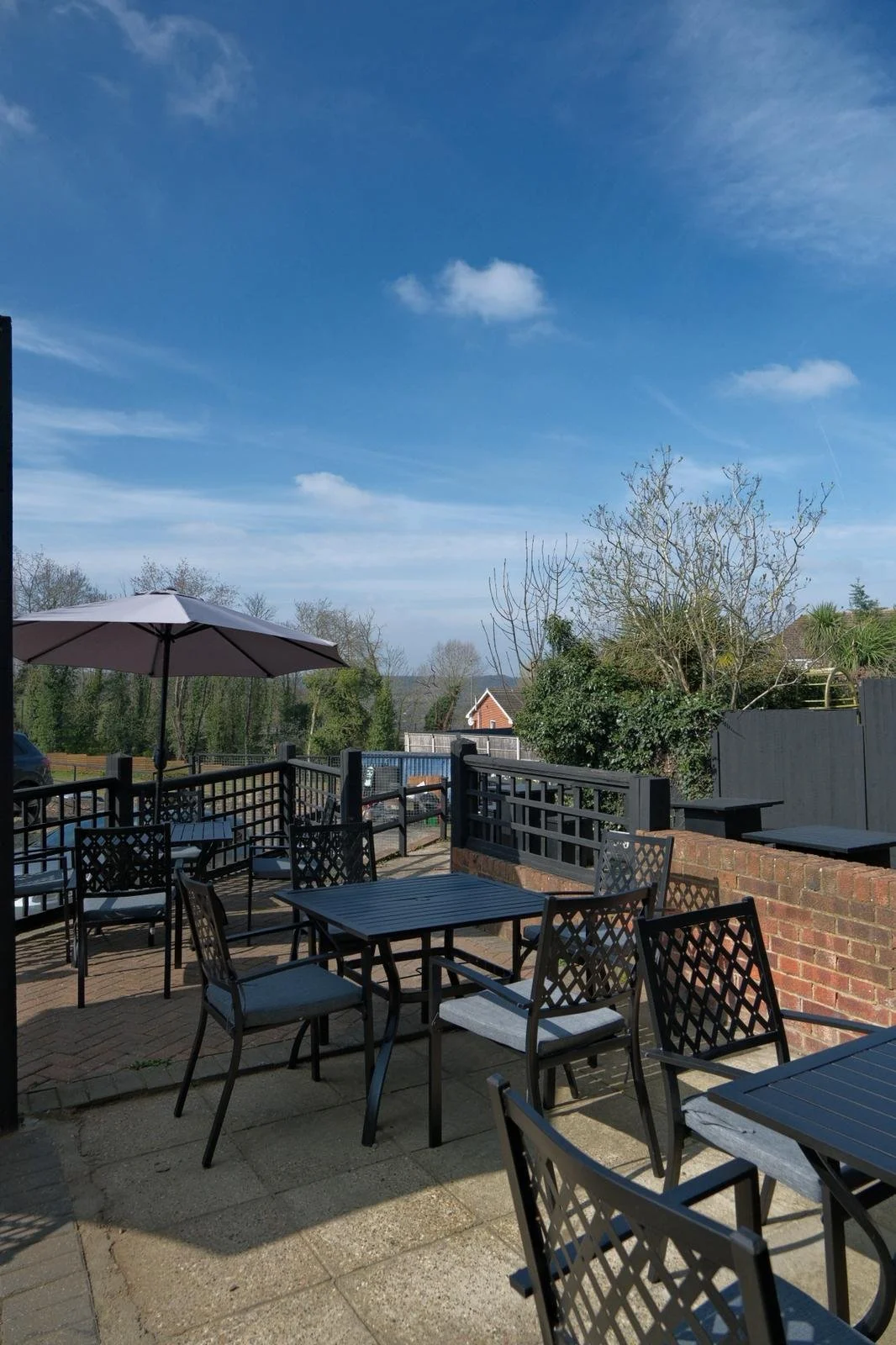 An outdoor patio with black metal tables and chairs, a large patio umbrella, surrounded by a black railing, with a view of a backyard and trees under a partly cloudy blue sky.