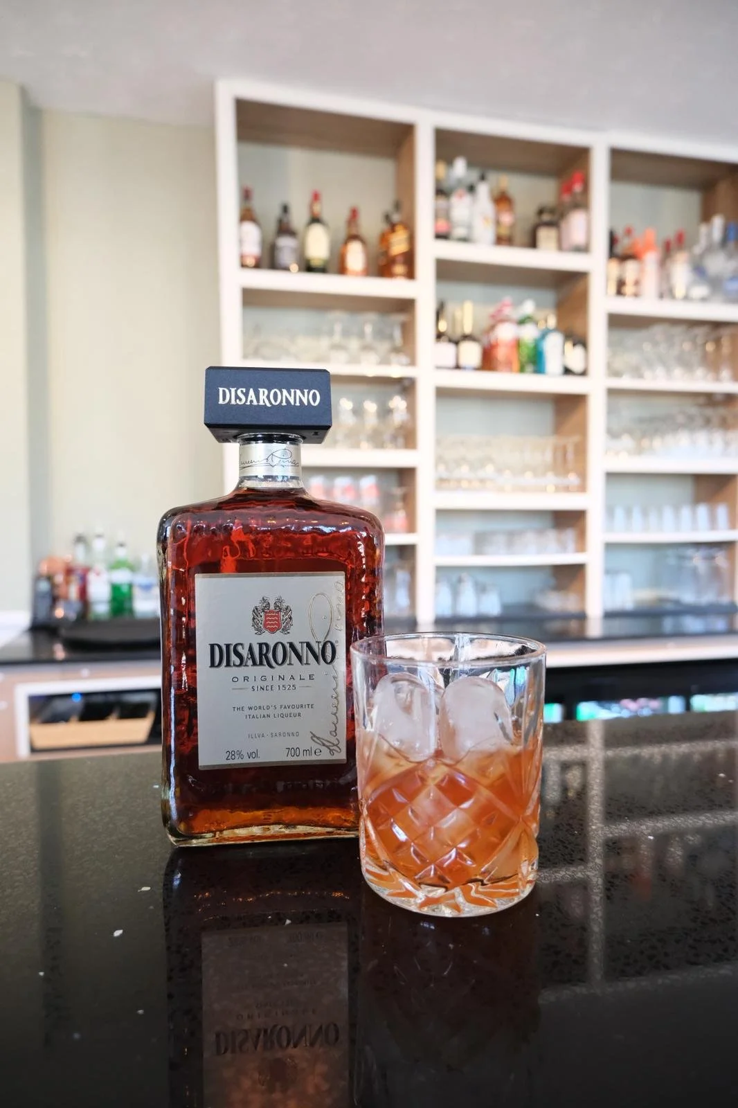 A bottle of Disaronno amaretto liqueur next to a glass with ice and a drink, on a bar counter with shelves of liquor bottles and glassware in the background.