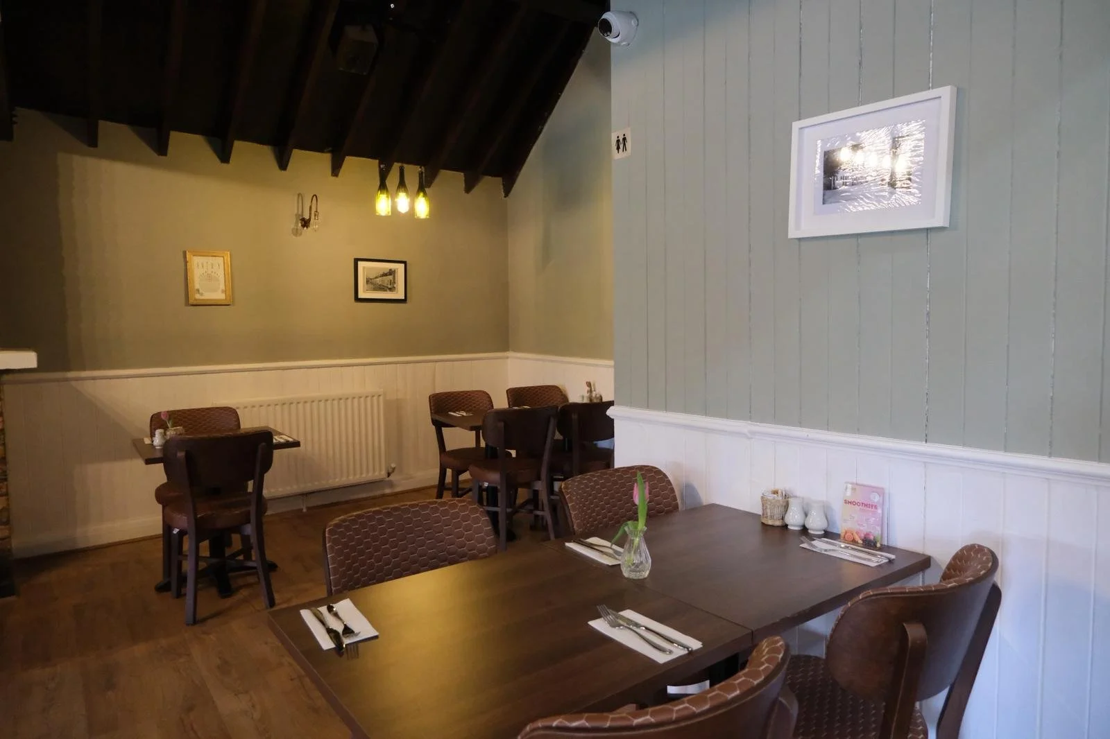 Interior of a cozy restaurant with wooden tables and chairs, some tables set with napkins and cutlery, featuring framed pictures on the walls, a green painted wall, and wooden ceiling beams.