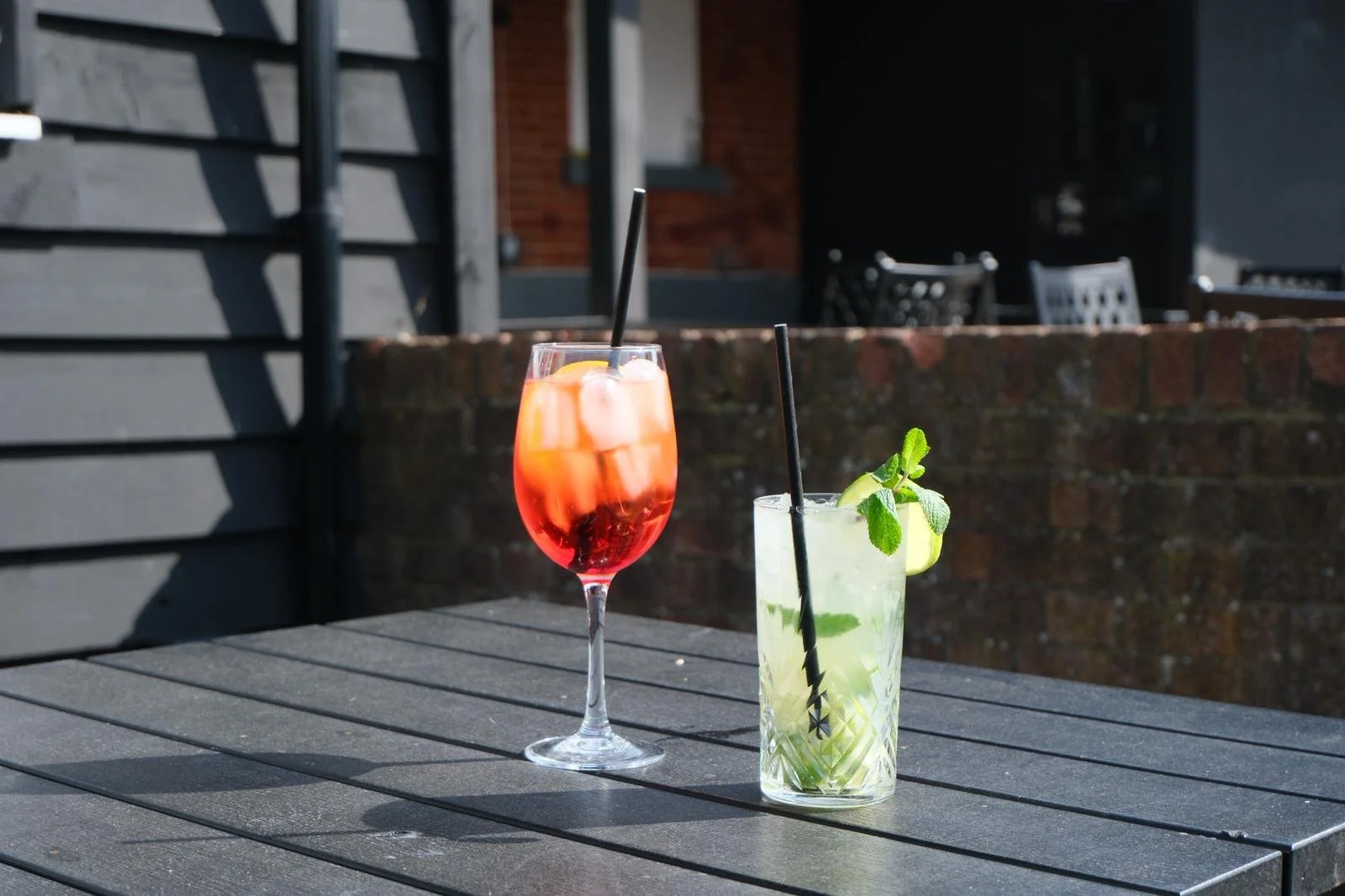 Two cocktails on a black wooden table outdoors, one a red tropical drink with ice in a tall glass, the other a light green mojito garnished with mint and lime, with black straws.
