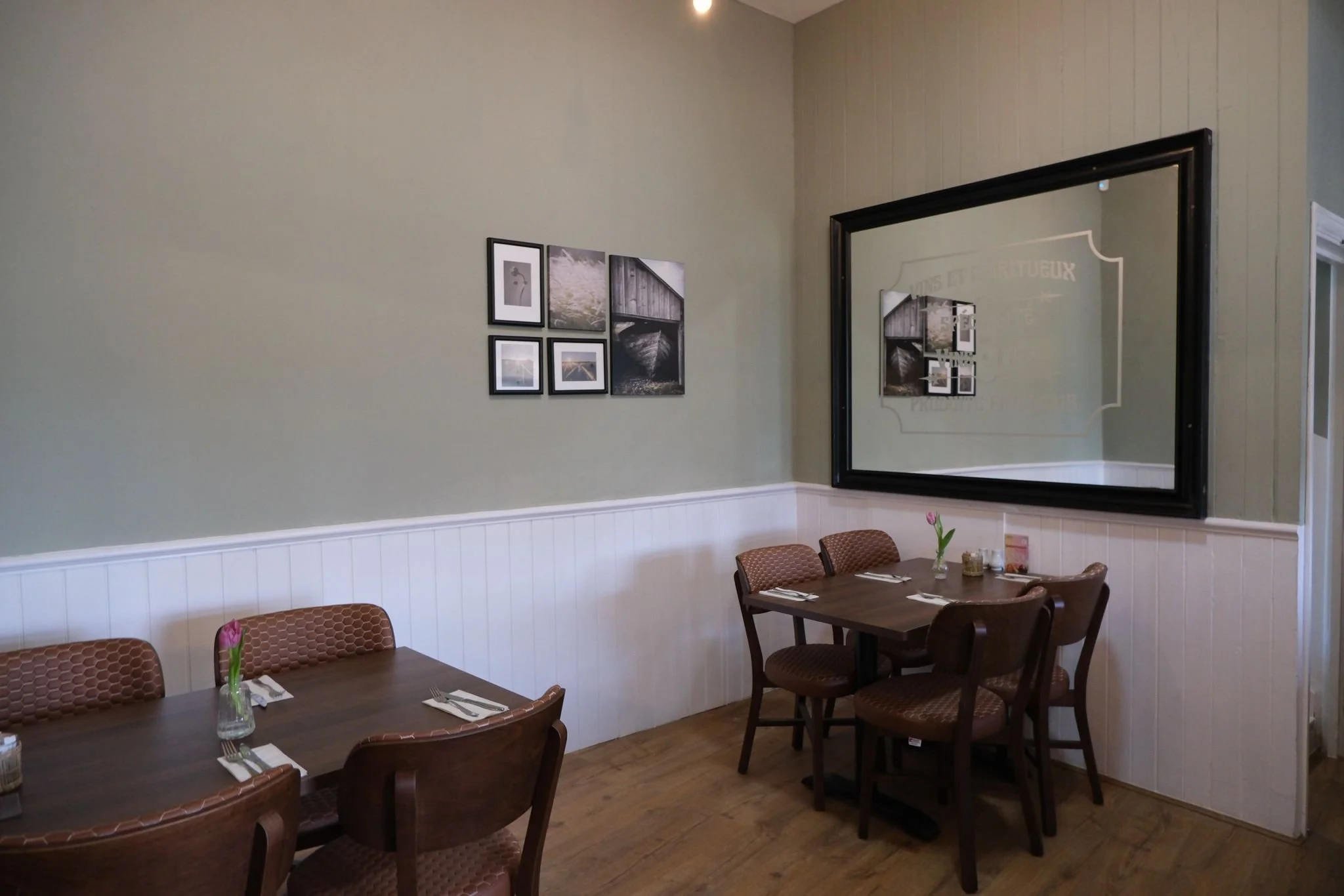 A cozy dining area with two wooden tables, each set with silverware, napkins, and a small vase with a pink tulip. There are six wooden chairs with cushioned seats around the tables. The walls are painted light green with white wainscoting, and a larg