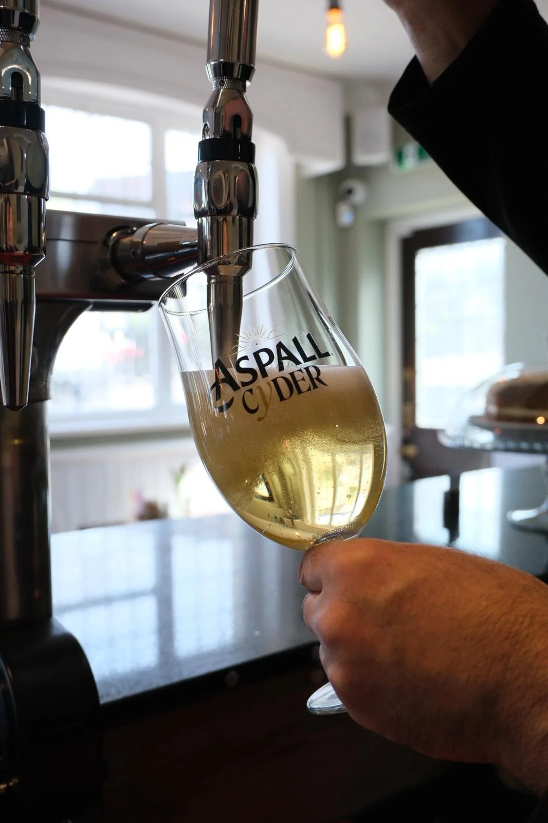 A hand holding a wine glass being filled with beer from a tap at a bar or restaurant, with a background of windows and interior