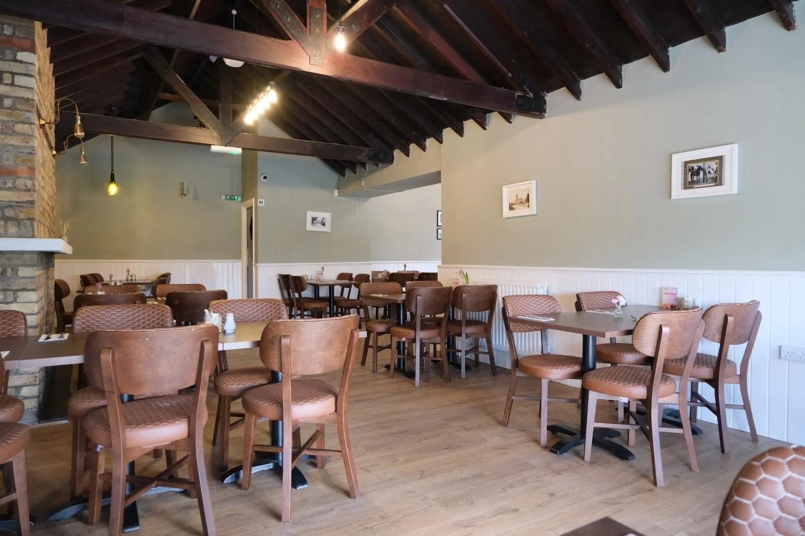 Empty restaurant with wooden tables and chairs, framed pictures on the wall, wood and brick accents, and a high ceiling with exposed beams.