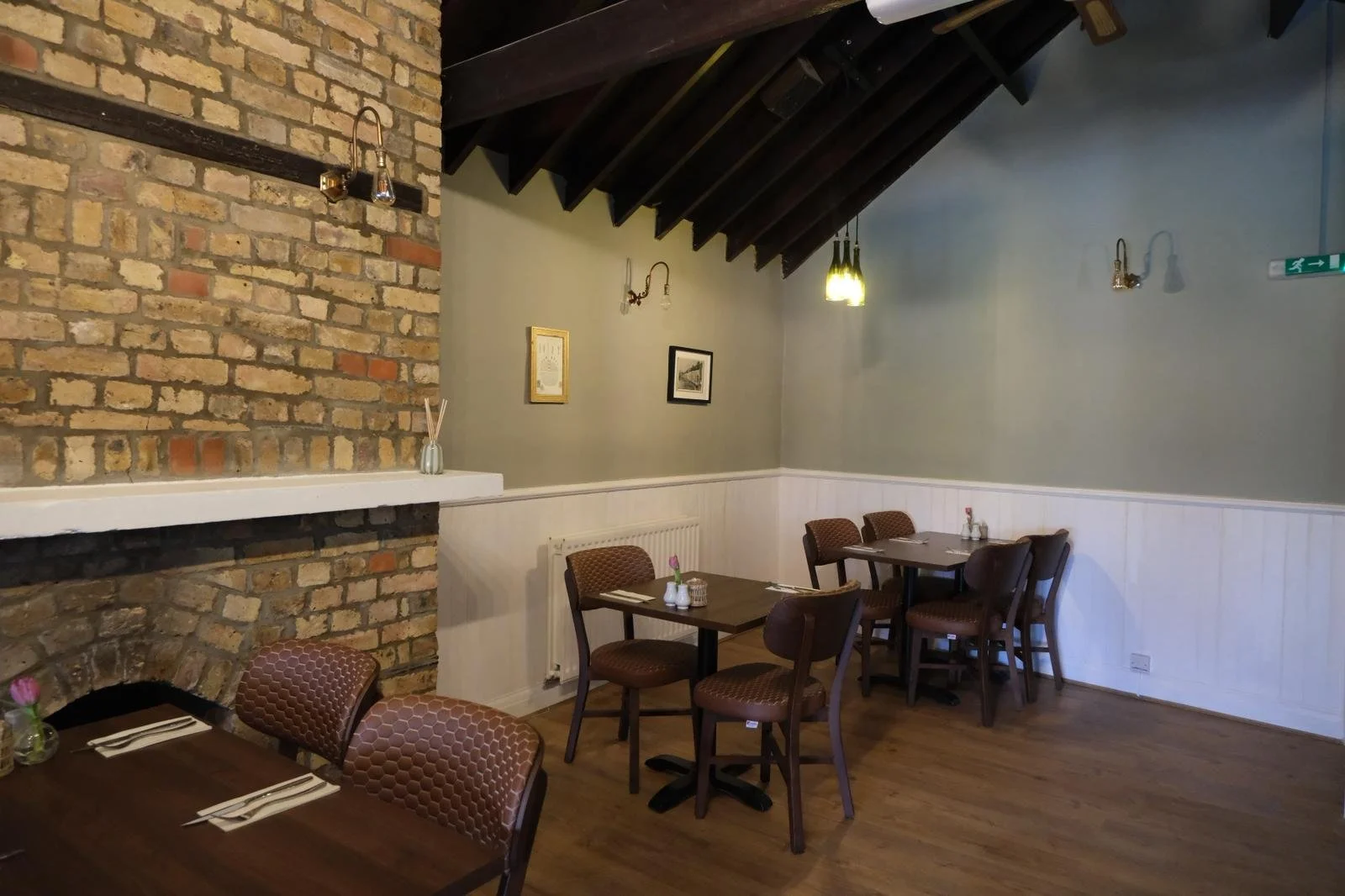 Interior of a cozy restaurant with exposed brick wall, dark wooden beams, framed artwork, and small tables set with napkins and flowers.