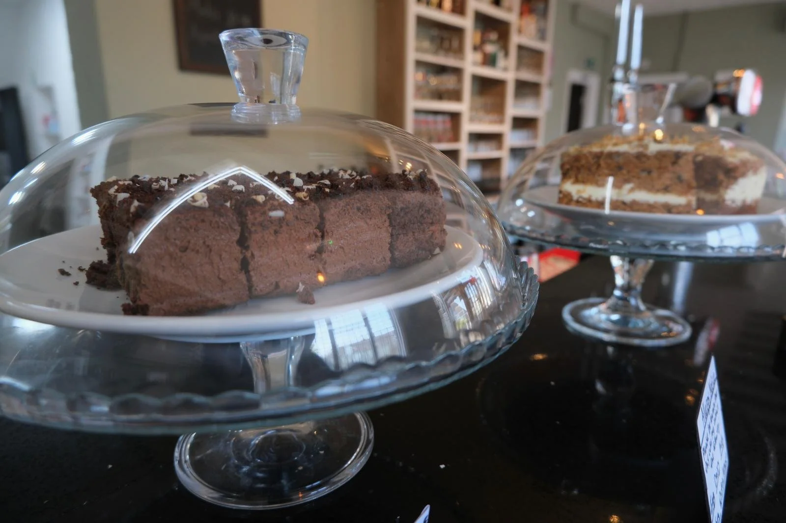 Two slices of cake on glass cake stands with covers. The front slice is chocolate with a chocolate topping, and the back slice appears to be a layered cake with cream and chocolate toppings. They are displayed on a dark surface, possibly in a bakery 