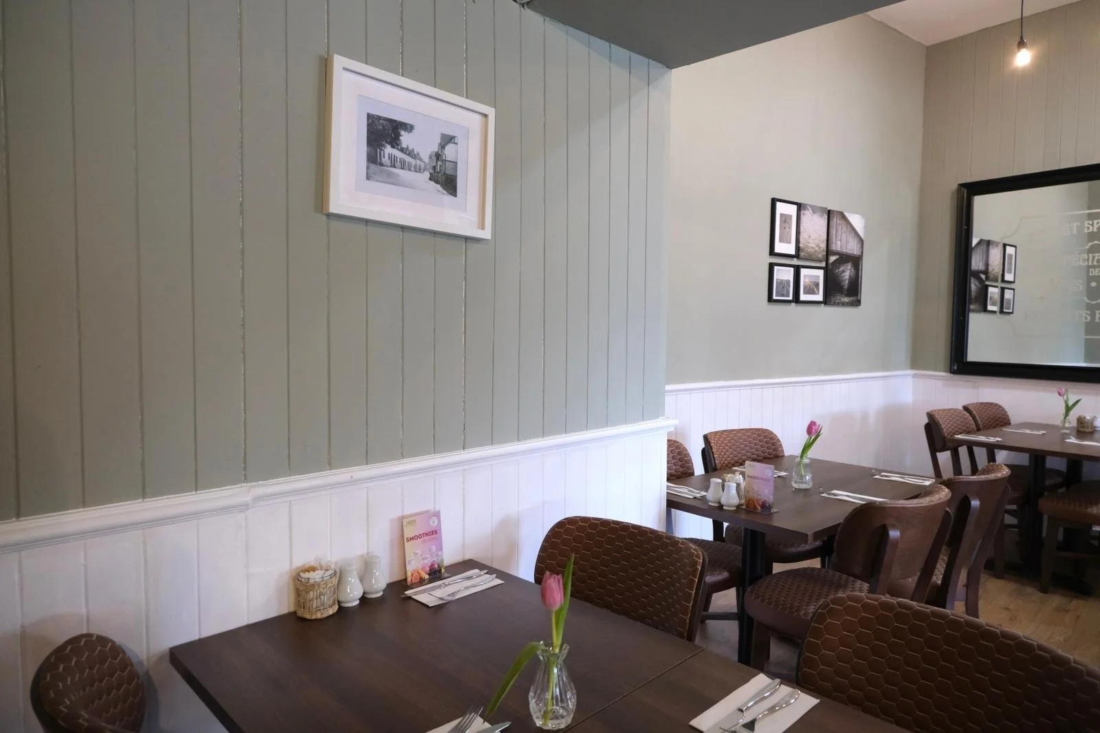 Cozy cafe with wooden tables, patterned chairs, pink tulips in glass vases, framed black and white photos on walls, and set tableware for dining.