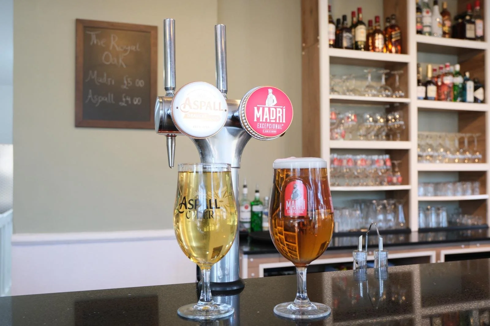 Two glasses of beer on a bar counter, one light-colored and one darker, with beer taps behind them in a bar with shelves of bottles in the background.