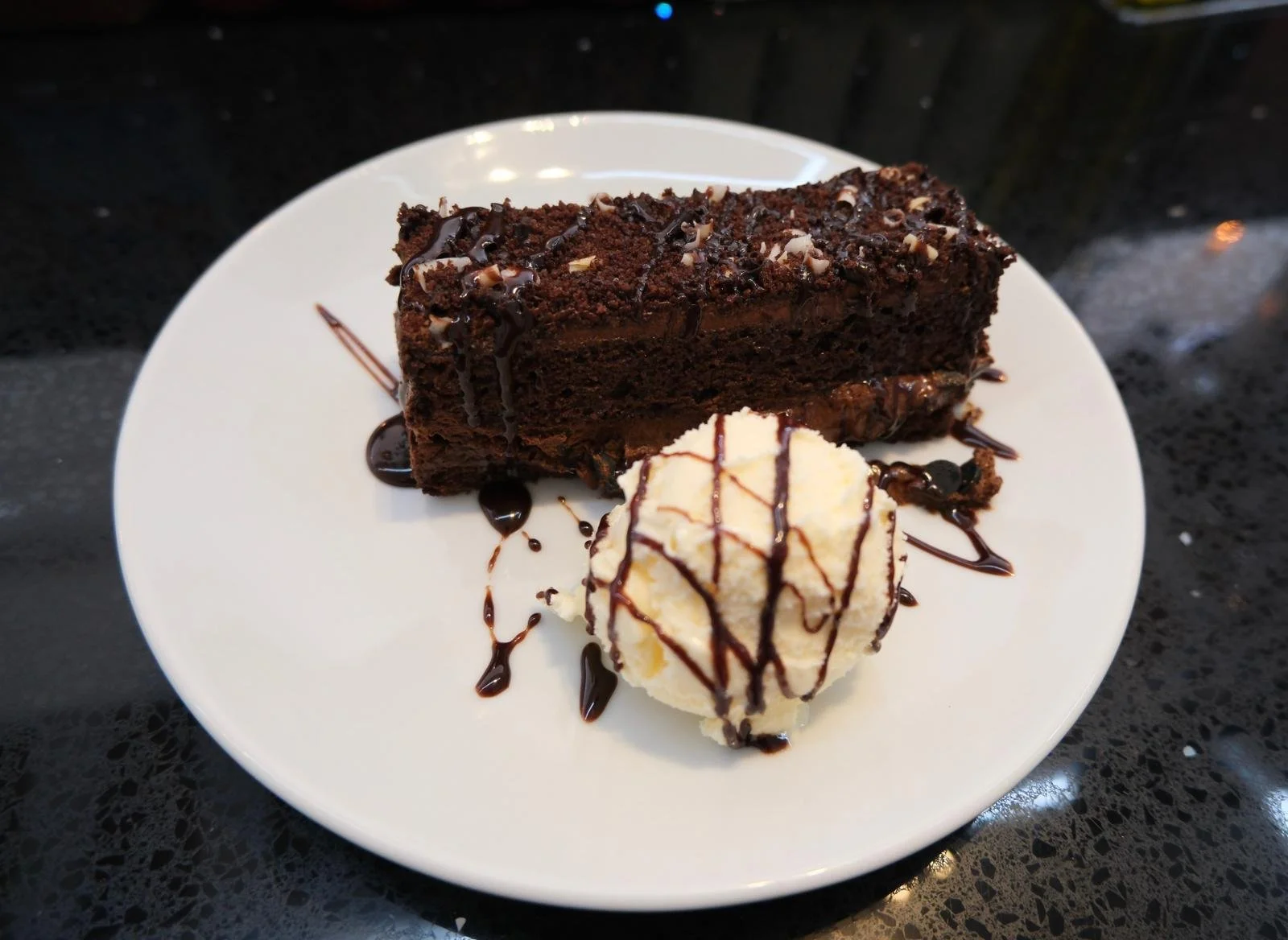 Chocolate cake with chocolate drizzle and a scoop of vanilla ice cream topped with chocolate syrup on a white plate.