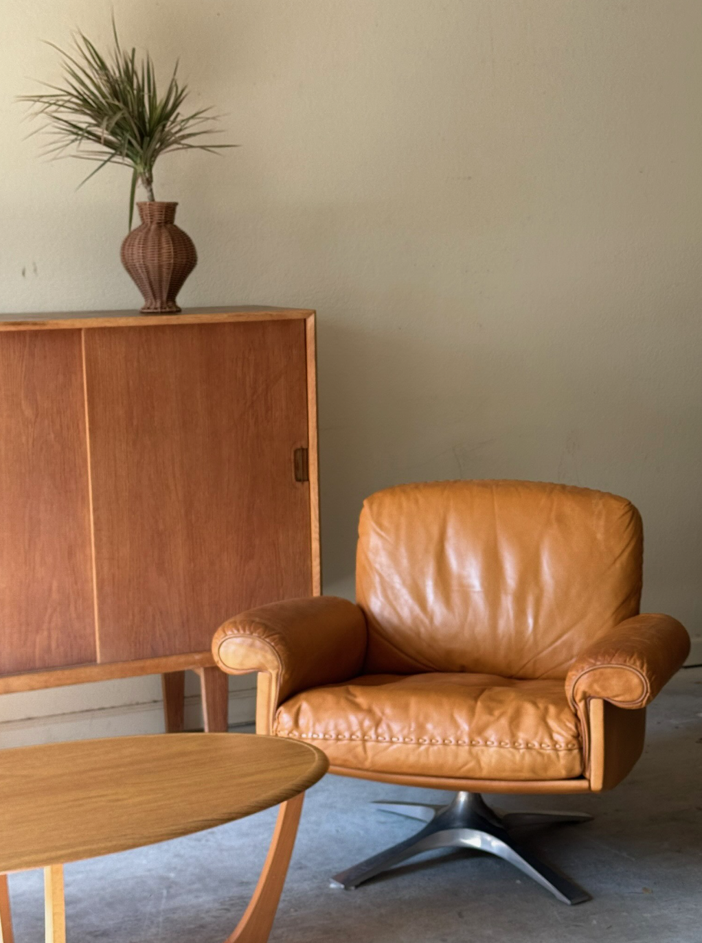 A mid-century modern leather armchair in front of a wooden sideboard with a decorative vase and plant on top.