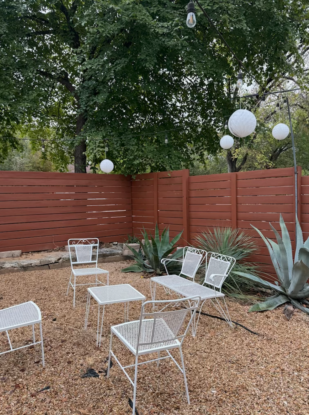 An outdoor backyard patio with white metal chairs and a small table, surrounded by desert plants, with a red wooden fence and string lights with paper lanterns hanging from trees.