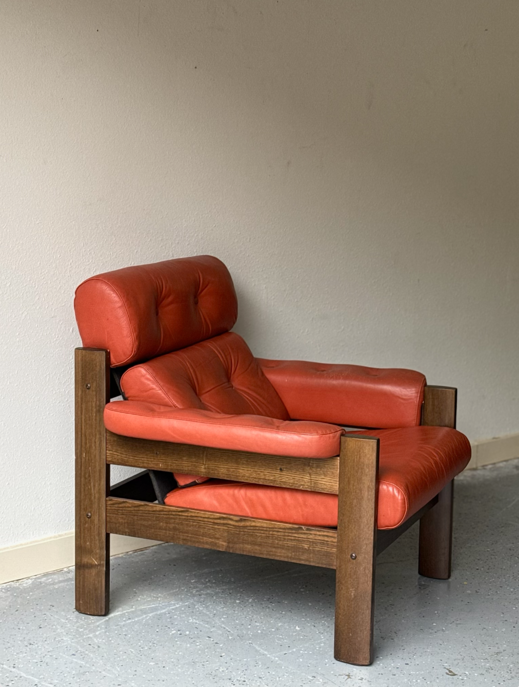 Red leather armchair with a wooden frame next to a beige wall on a gray speckled floor.