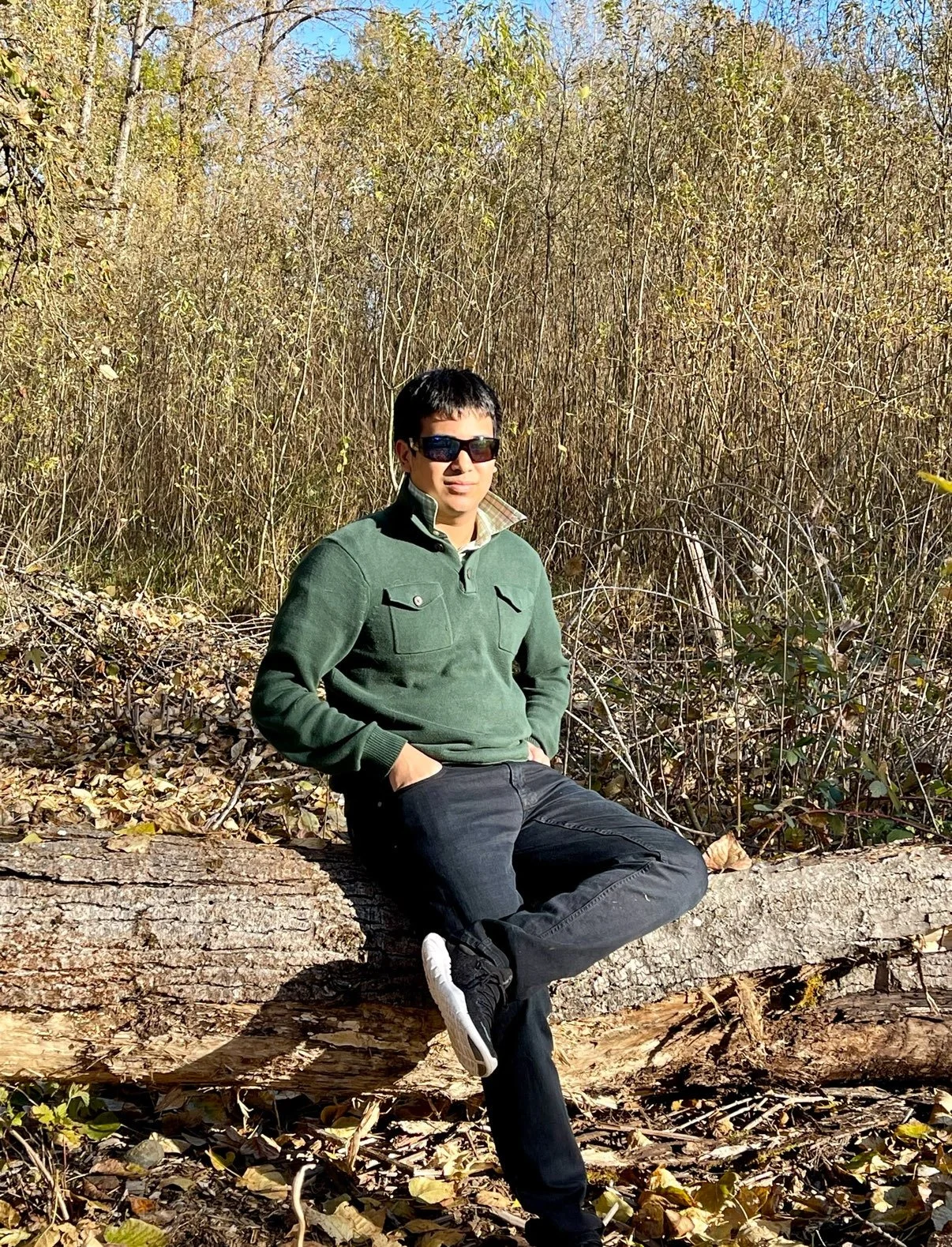 A man in sunglasses and a green jacket sitting on a fallen tree trunk in a wooded area with bare trees and fallen leaves.