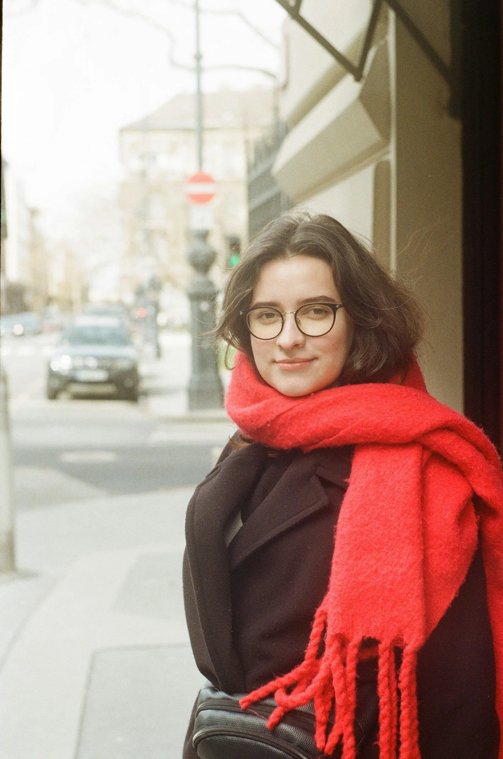 A woman with shoulder-length wavy dark hair, wearing glasses, a black coat, and a red scarf, standing outdoors on a city street with cars and a stop sign in the background.