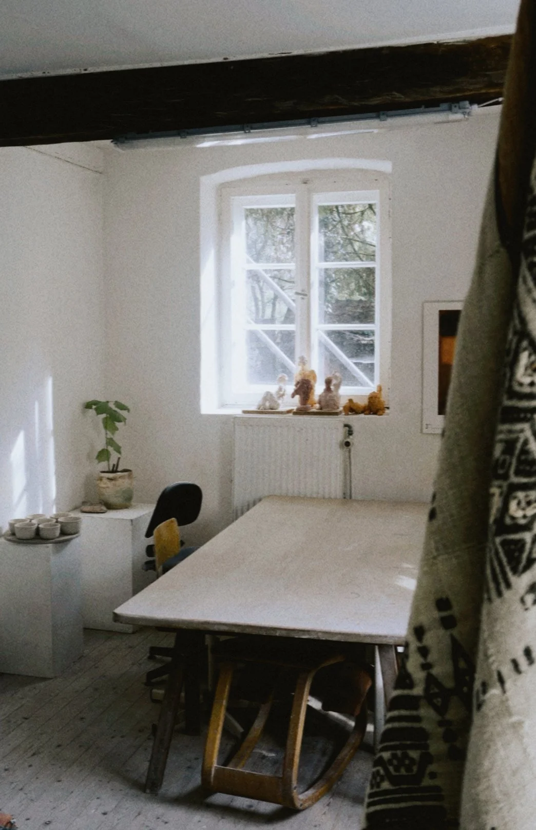 A room with a large window letting in natural light, decorated with teddy bears on the windowsill, a white table with chairs underneath, a plant in a pot on a small white table, and a black chair. The room has light-colored walls, wooden floor, and exposed wooden beam on the ceiling.