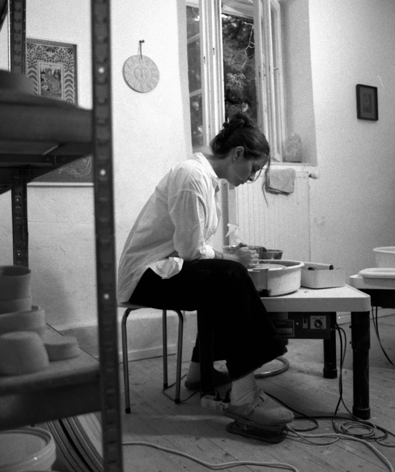 A woman in a white shirt and black pants sitting on a stool, working on pottery with tools, in a room with a window and shelves.