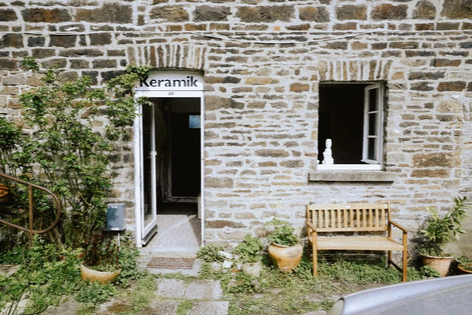 Stone building with an open door labeled 'Keramik', a window with a white figurine, surrounded by potted plants and a wooden bench.
