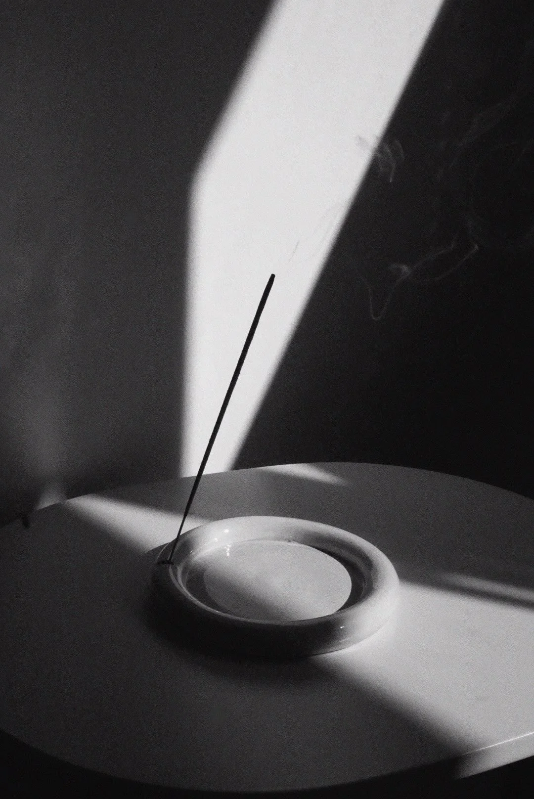 A bowl with a stick, possibly incense, is placed on a circular plate. A white rectangular object, like a door or panel, is behind the setup, casting shadows and creating a monochrome scene.