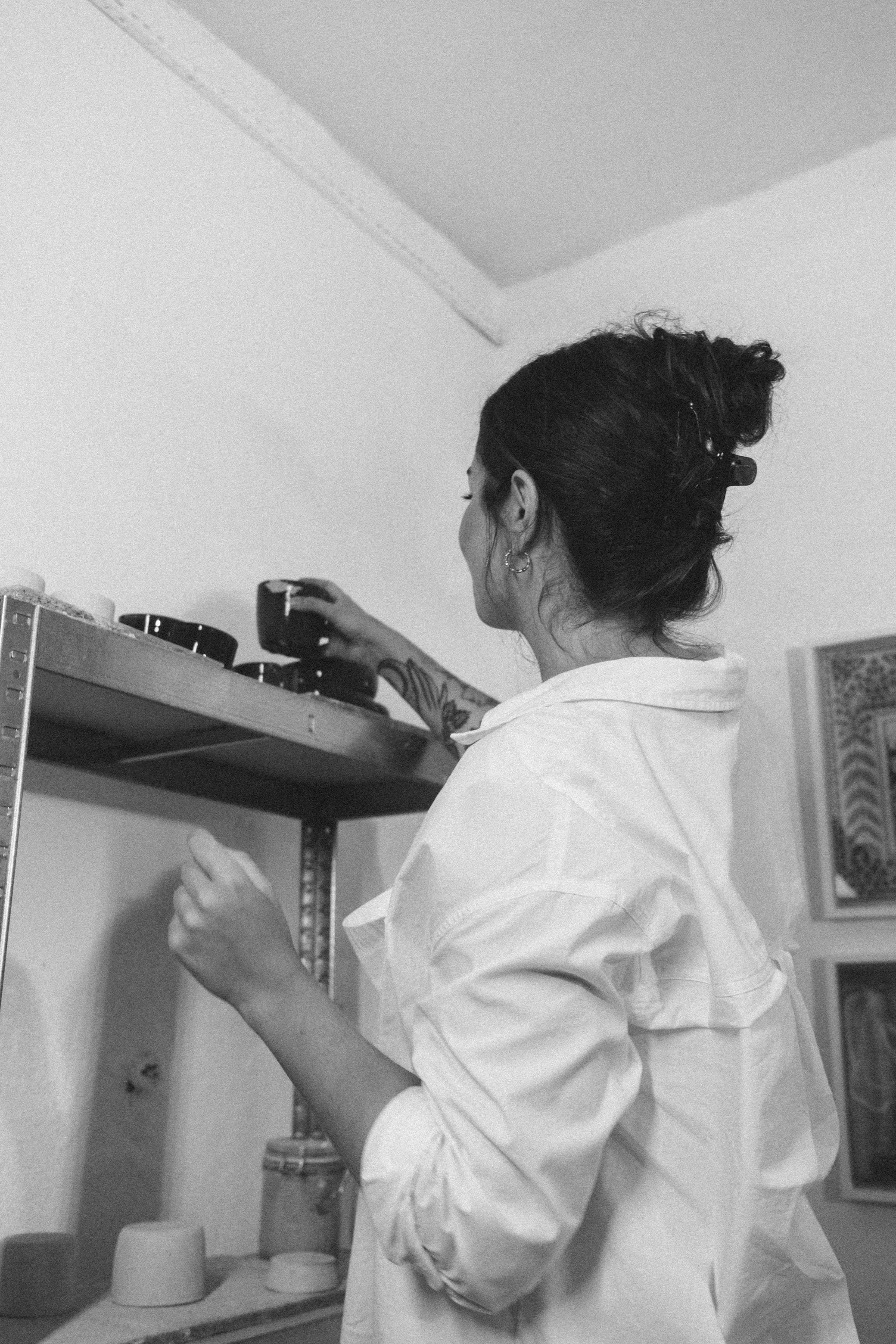 A woman with dark hair in a bun, wearing a white shirt, is reaching for a small black cup on a metal shelf in a room.