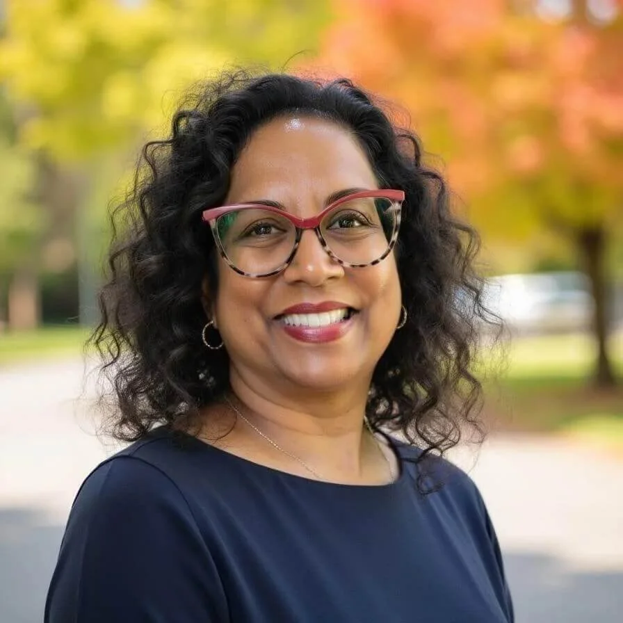 Image of a woman with black, shoulder length curly hair, and tortoise shell glasses with a red top rim.  She is wearing a navy blue blouse.  This is a headshot.