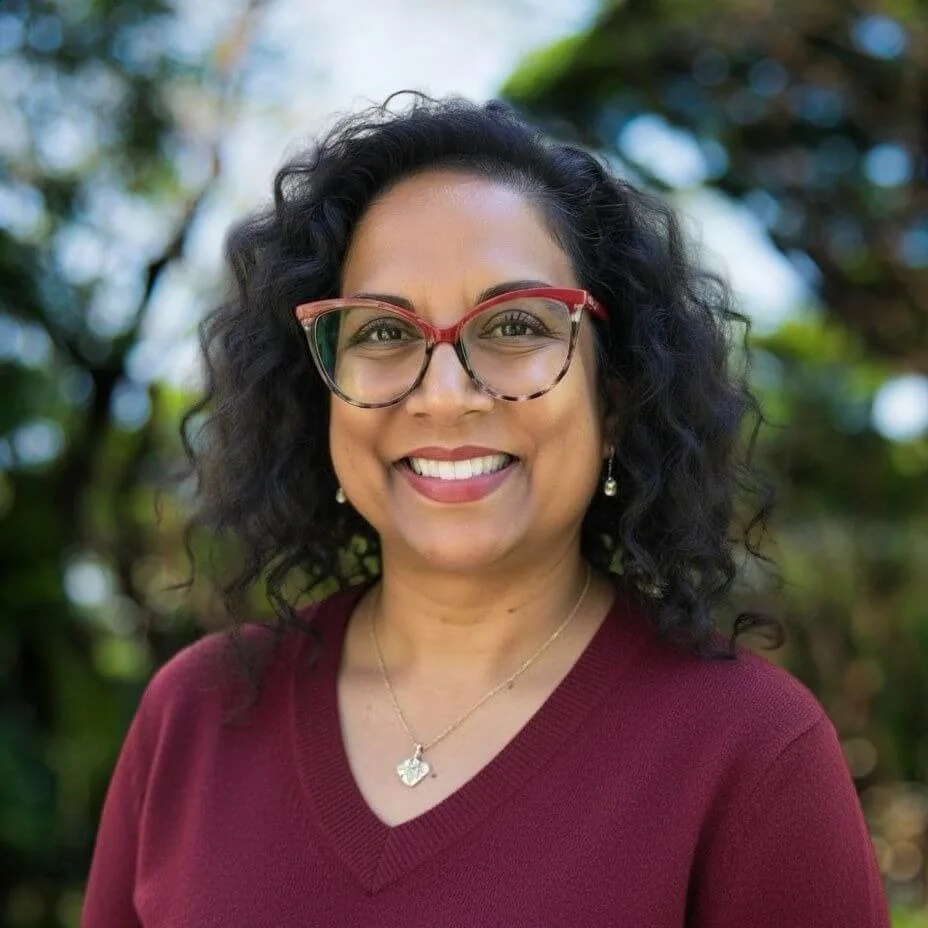 Picture of a woman with black curly hair, red rimmed glasses and a wine colored sweater.  This is a headshot.