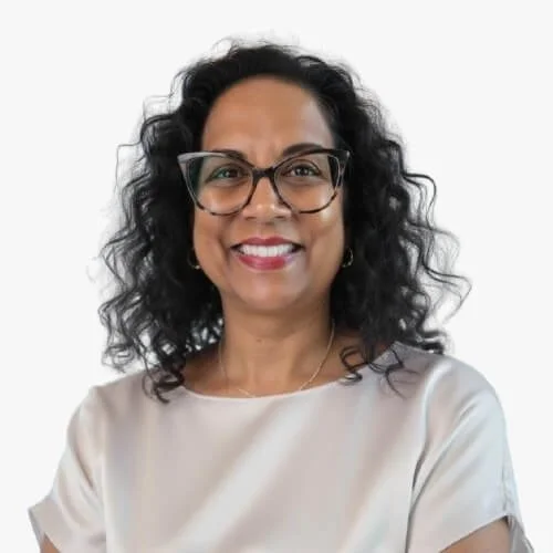 Picture of a woman with shoulder length, black curly hair.  She has tortoise shell glasses and a cream blouse.  The background is pink.  This is a headshot.