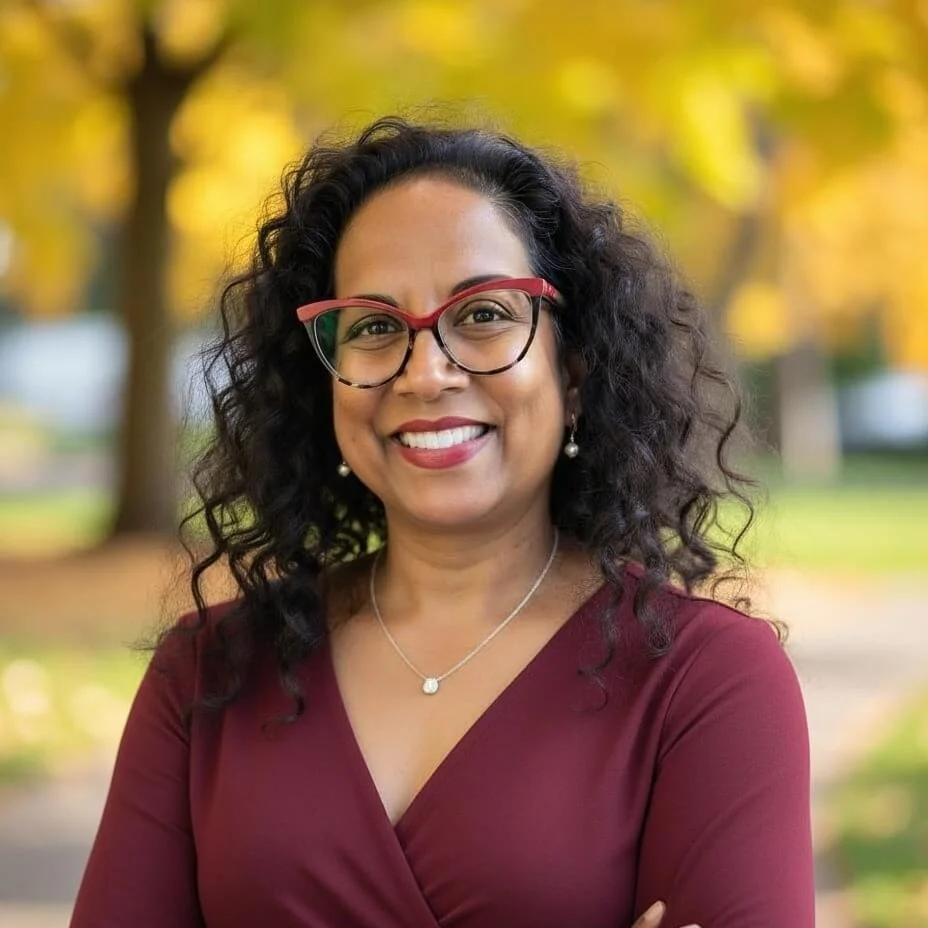 Woman with shoulder length dark curly hair and tortoise shell glasses with a red upper rim.  She is wearing a wine blouse.  This is a heashot.