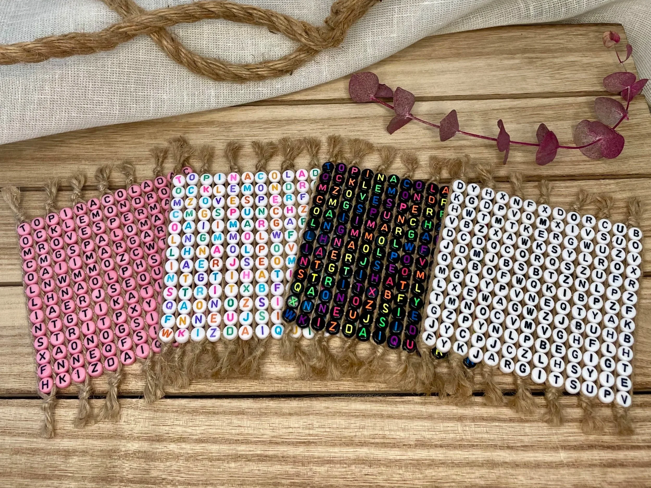 Four beaded word search coasters arranged on a wooden surface, with a piece of rope and a sprig of purple leaves nearby.