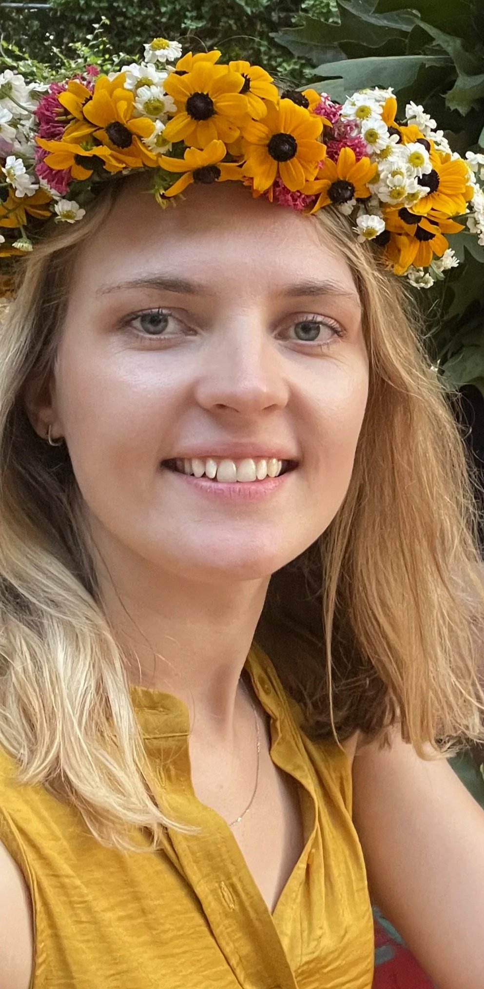 A young woman with blonde hair and blue eyes wearing a yellow top and a flower crown made of yellow, white, and pink flowers, smiling outdoors.