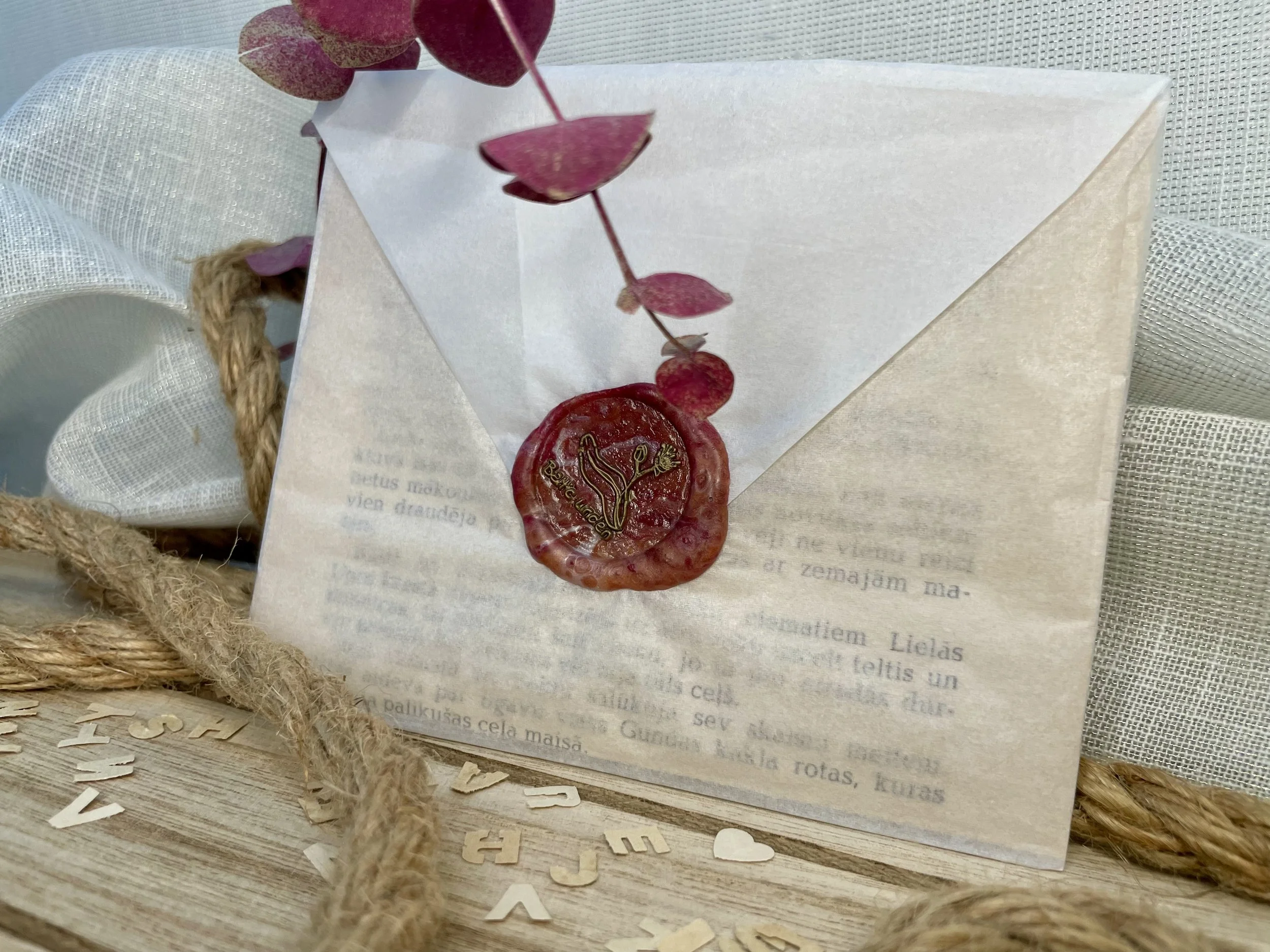 A white envelope with a wax seal and a small branch with pink leaves, resting on a wooden surface decorated with scattered letter cutouts and a piece of white fabric.