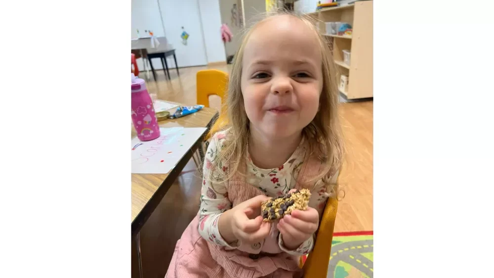 Scarlett's Fund. A young girl with long blonde hair and a floral shirt is sitting at a table, holding a cookie, and making a playful face. There are colorful items on the table and a classroom setting in the background.