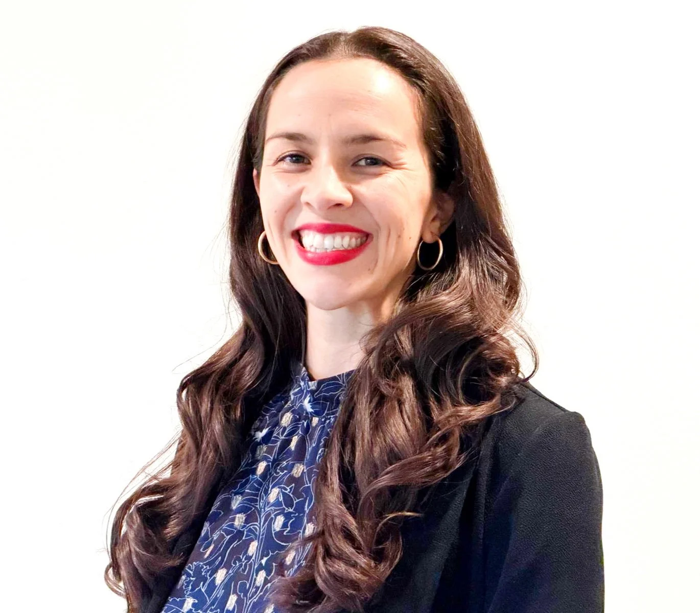 Iliana Reinhardt the treasurer and president of Reinhardt Consulting. A woman with long, curly brown hair, wearing a dark blazer and a patterned blouse, smiling with red lipstick, against a plain white background.