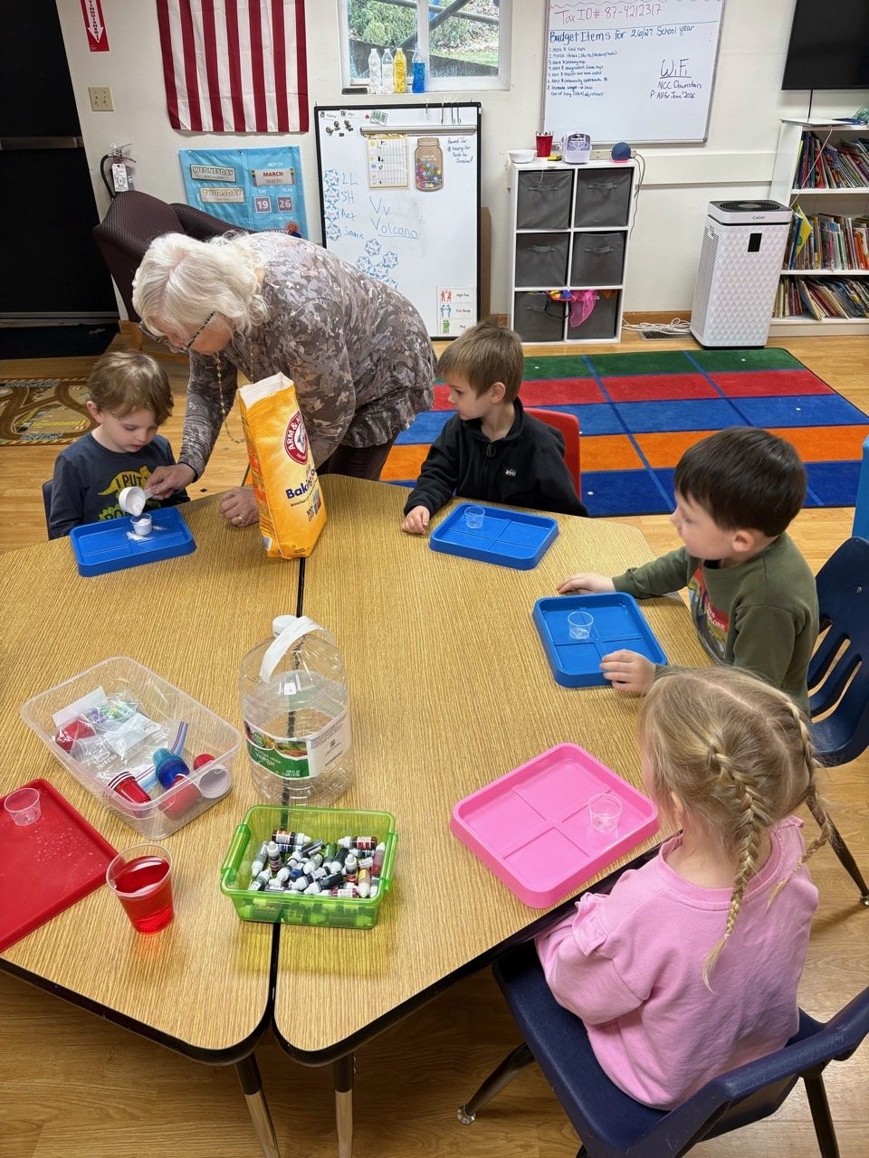 A classroom with four young children seated around a round table, engaged in an activity involving small plastic cups and various items. An adult woman is assisting a boy on the left. The table has containers of small objects, a water jug, and a red cup. The background includes a whiteboard, bookshelf, colorful carpet squares, and classroom decorations.