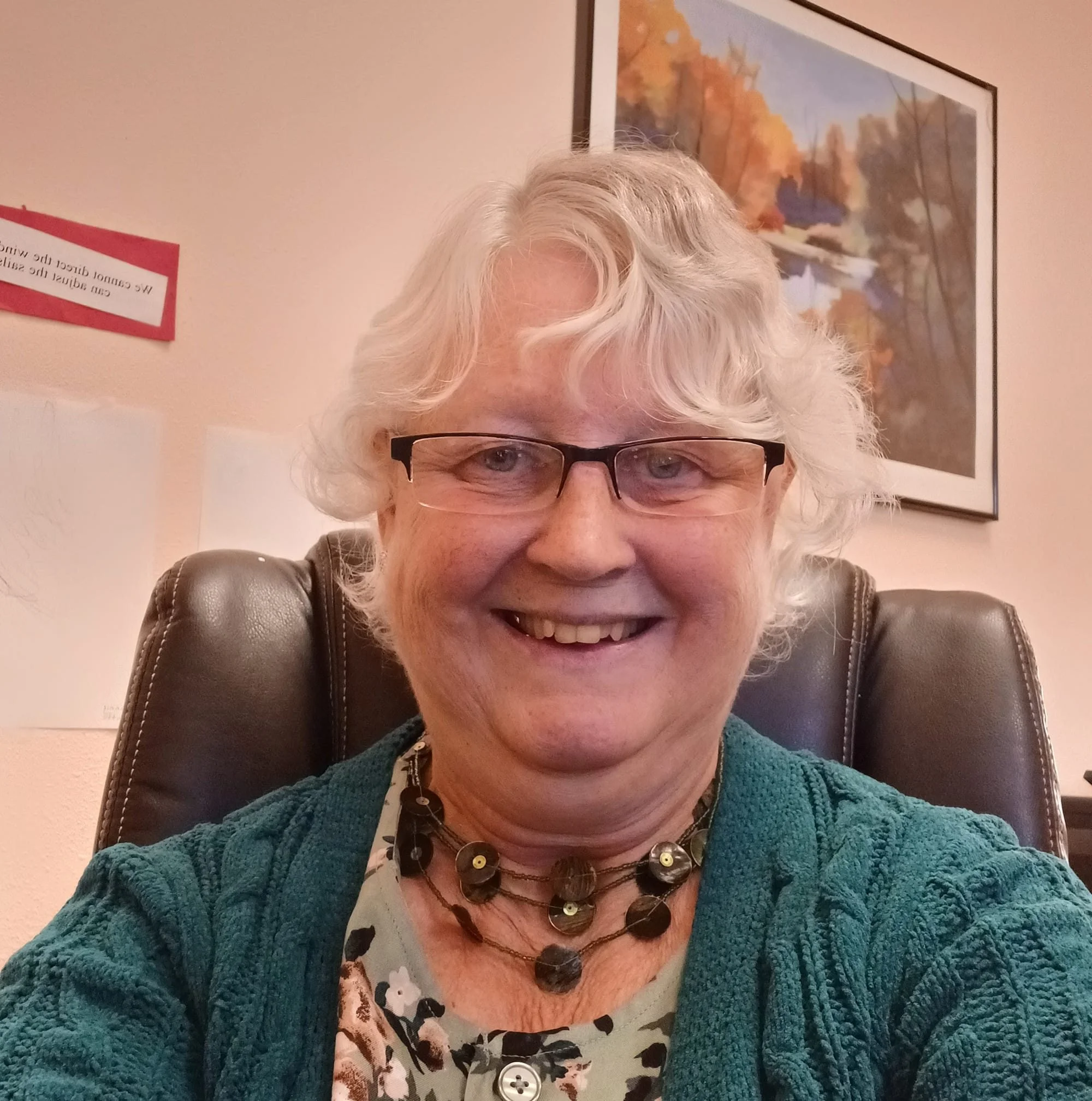Ginger Case a preschool teacher. A person with white hair, glasses, and a colorful necklace, wearing a teal sweater and patterned shirt, sitting on a brown leather chair in front of a painting of a river and autumn trees on a beige wall.
