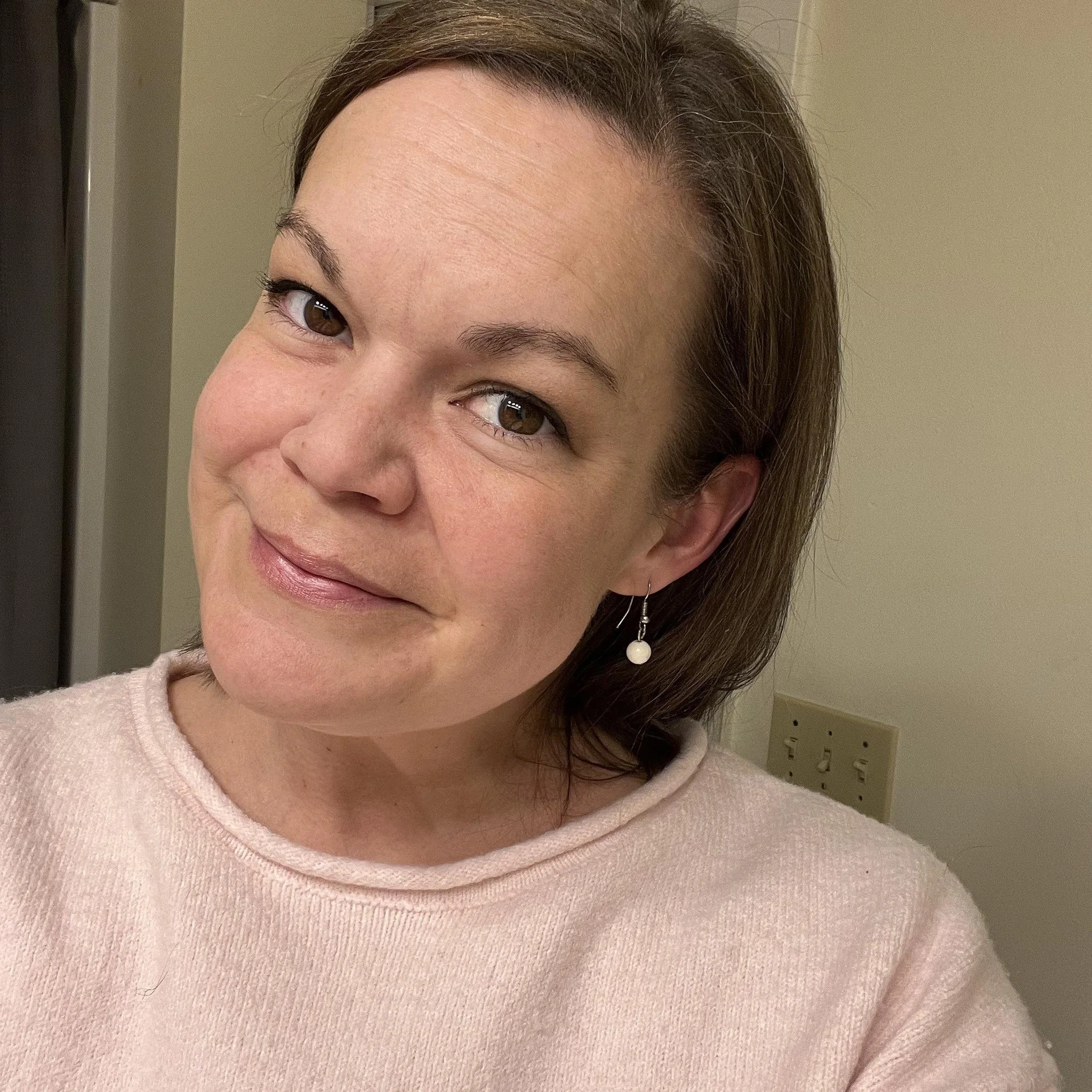 Alicia Charko a preschool teacher. Close-up of smiling woman with brown hair and hoop earring, wearing a soft pink sweater.