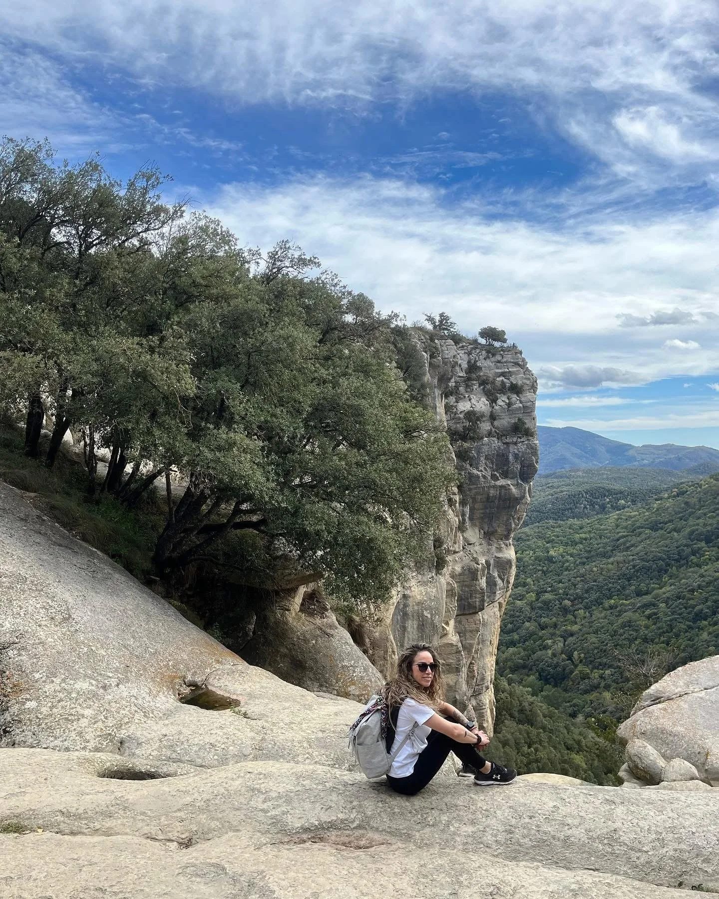 Mujer sentada sobre una roca en un paisaje montañoso con árboles y una formación rocosa grande y escarpada, cielo parcialmente soleado con nubes.