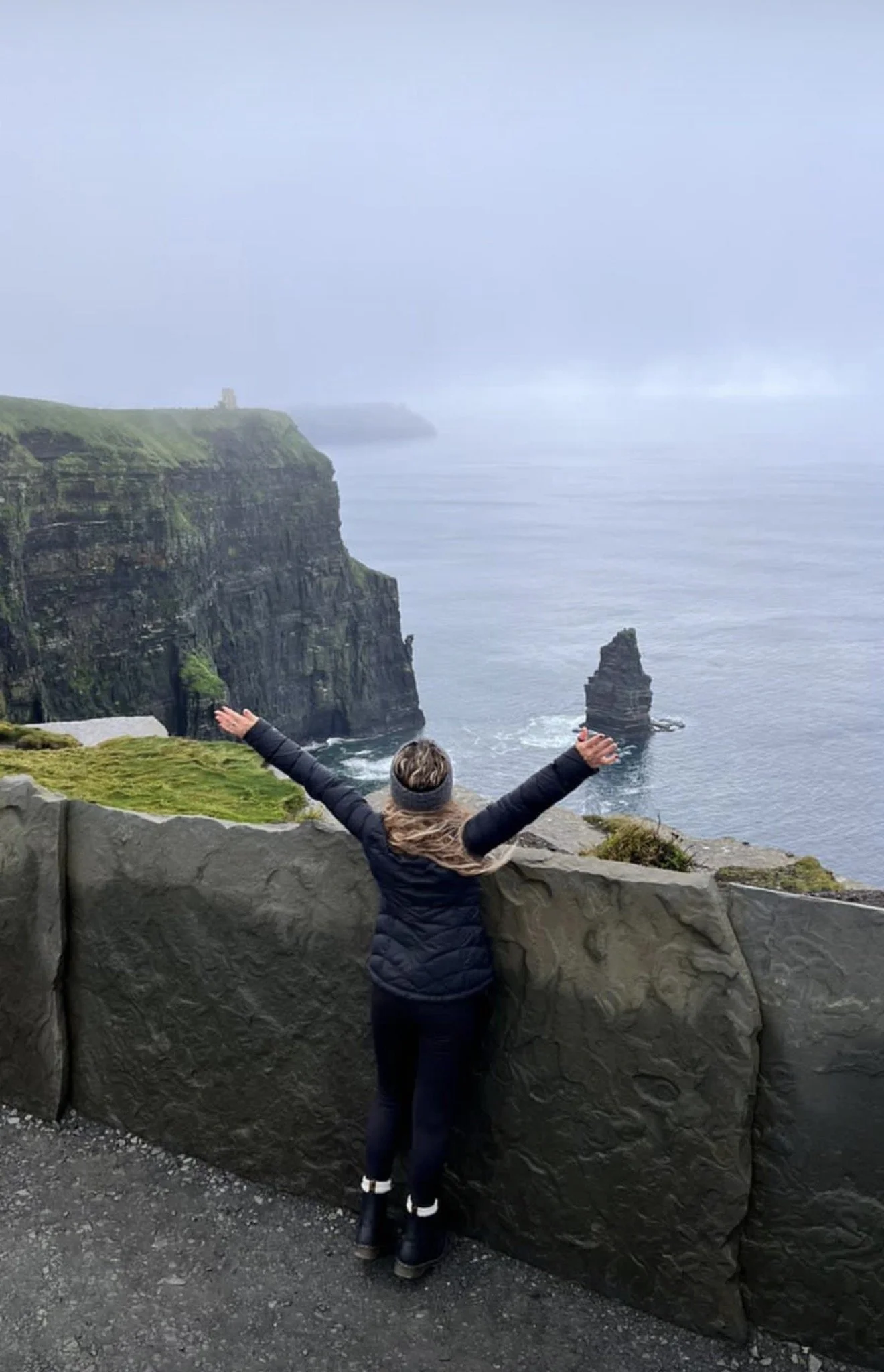 Una mujer de espaldas con los brazos abiertos, sintiendo la magia de viajar, mirando los acantilados y el mar en un día nublado en la Costa de Irlanda.