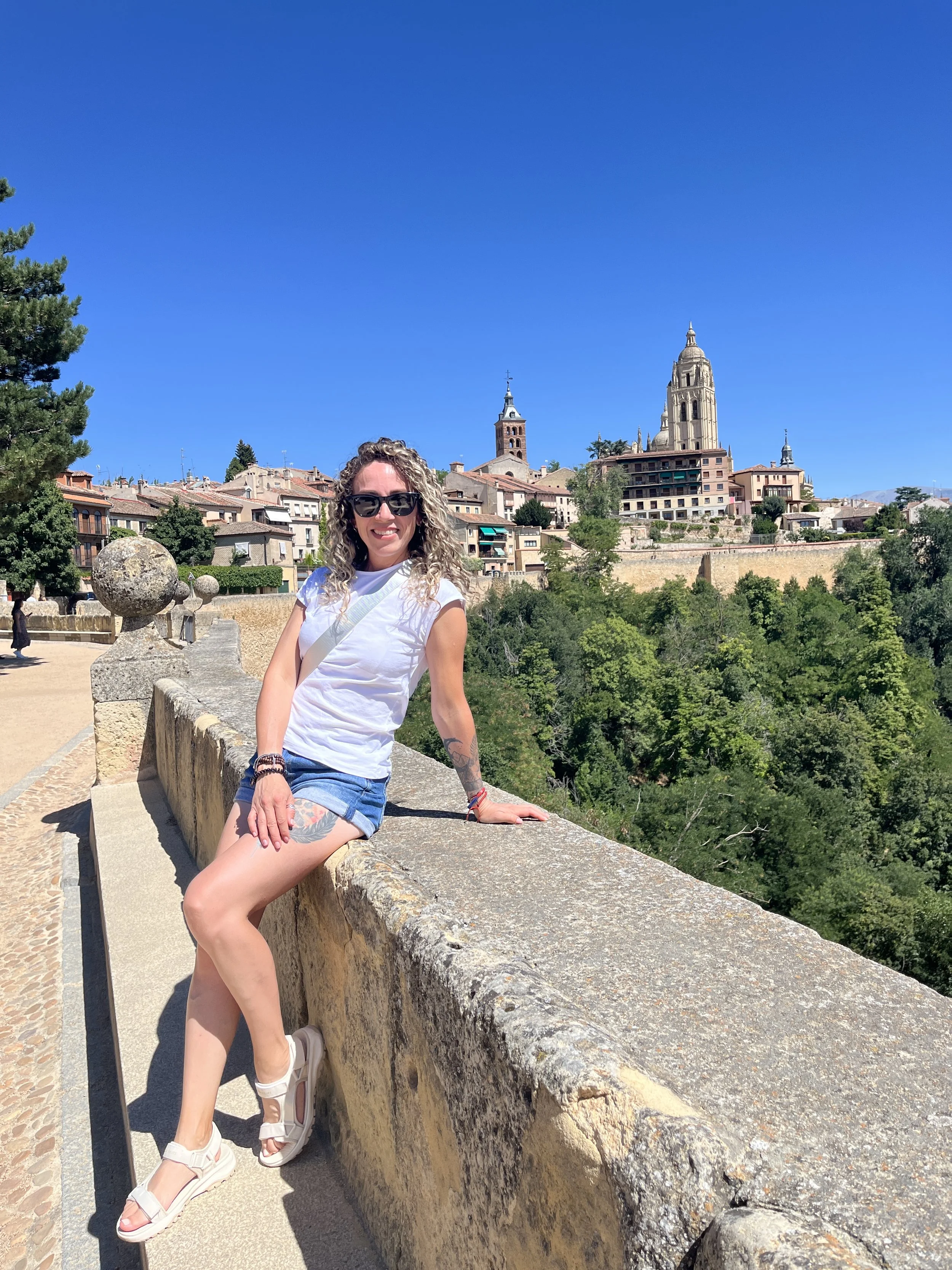 Una mujer con gafas, camiseta blanca y pantalones cortos de mezclilla, sentada sobre un muro de piedra, con la ciudad y la catedral en el fondo bajo un cielo azul claro.